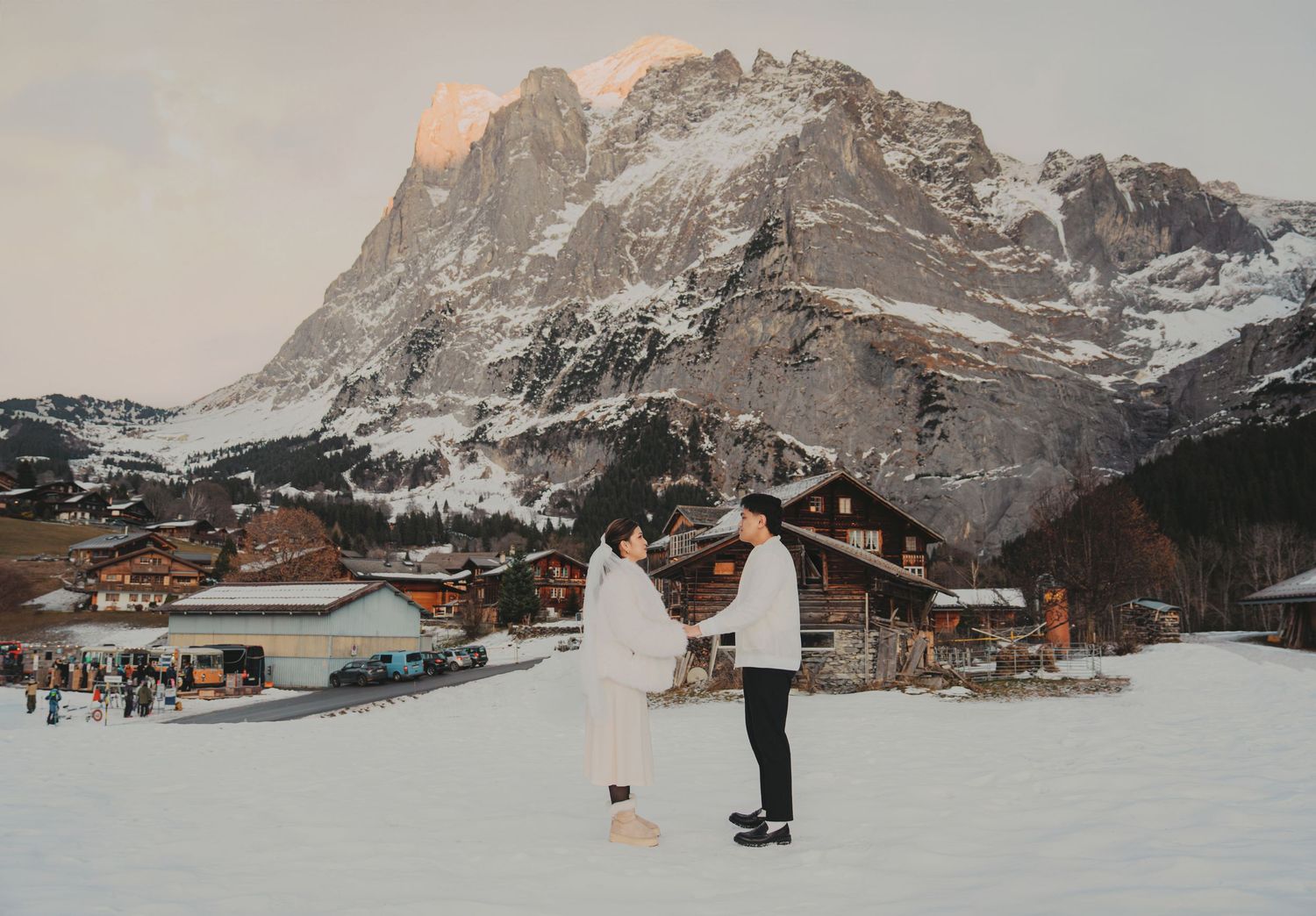 Couple holding hands in white outfits on snowy field with charming alpine village and mountain view.