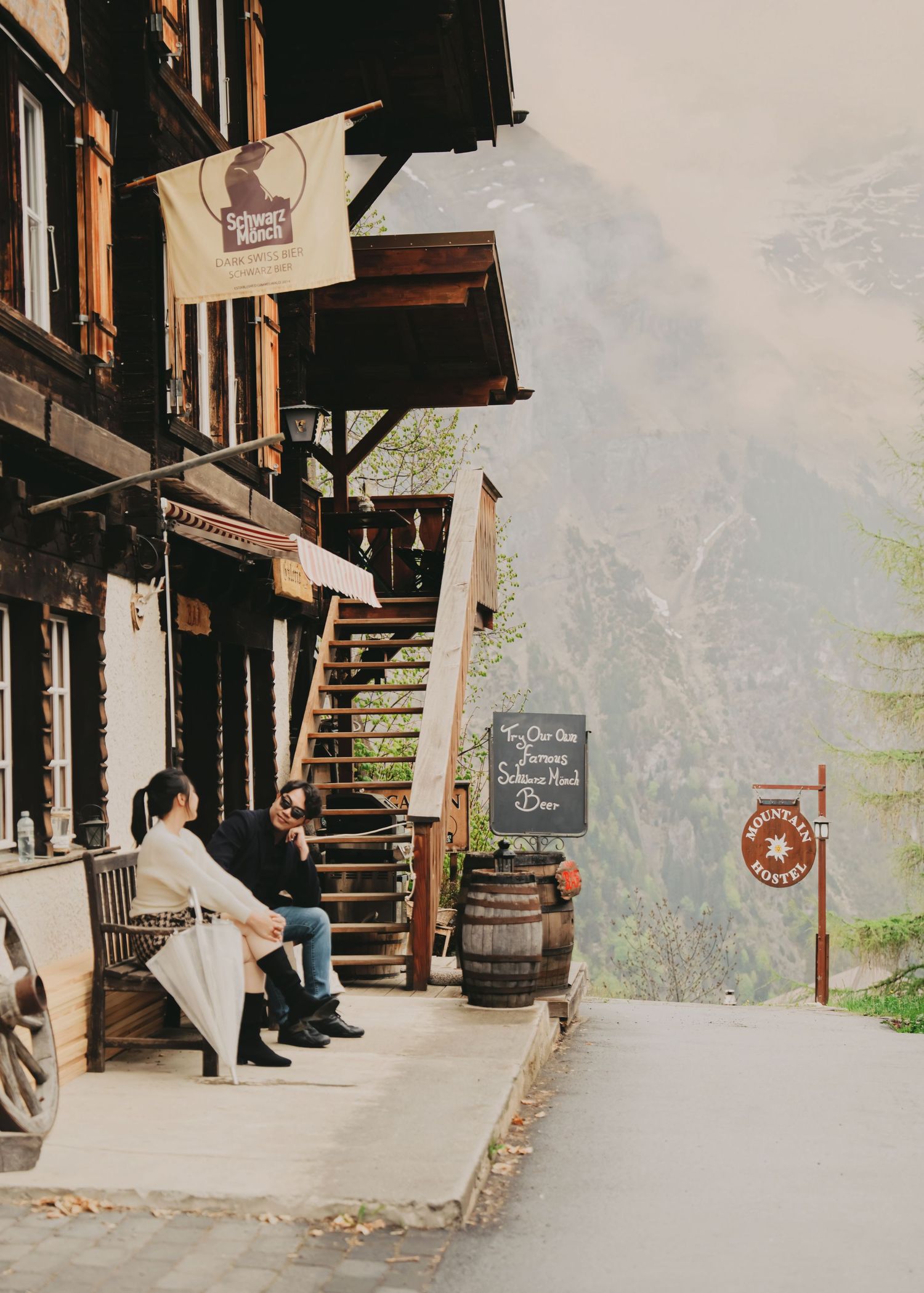 Couple resting on bench outside rustic alpine wooden building with mountain mist backdrop.