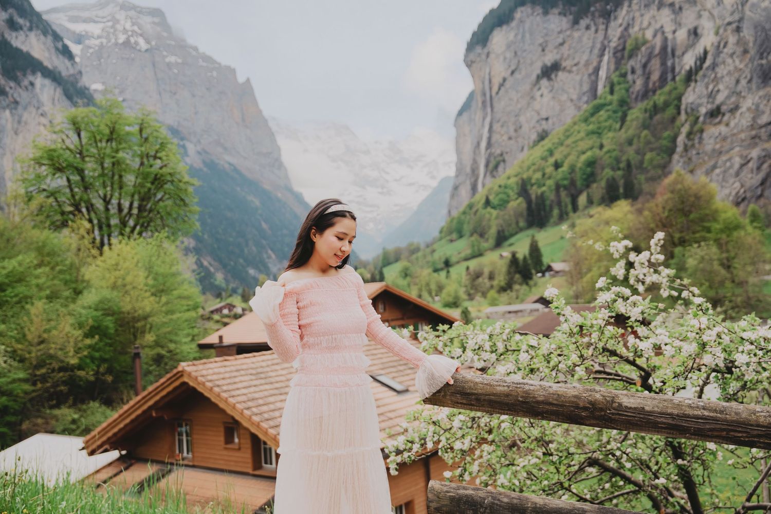 Woman in pink dress touching blooming cherry tree with Swiss Alps and Lauterbrunnen valley in background.
