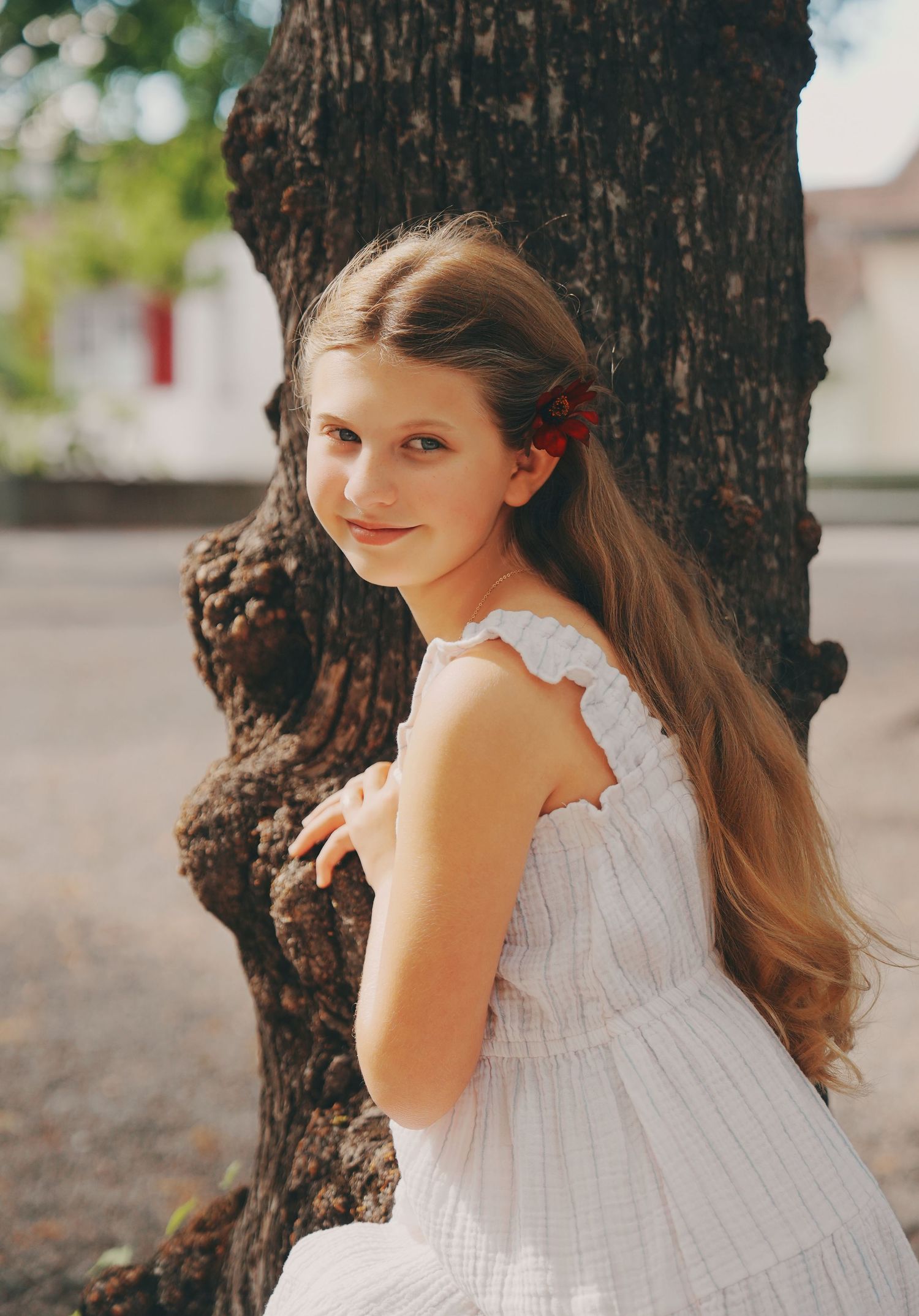 A smiling young girl with long hair in a white dress leans against a large tree trunk outdoors.