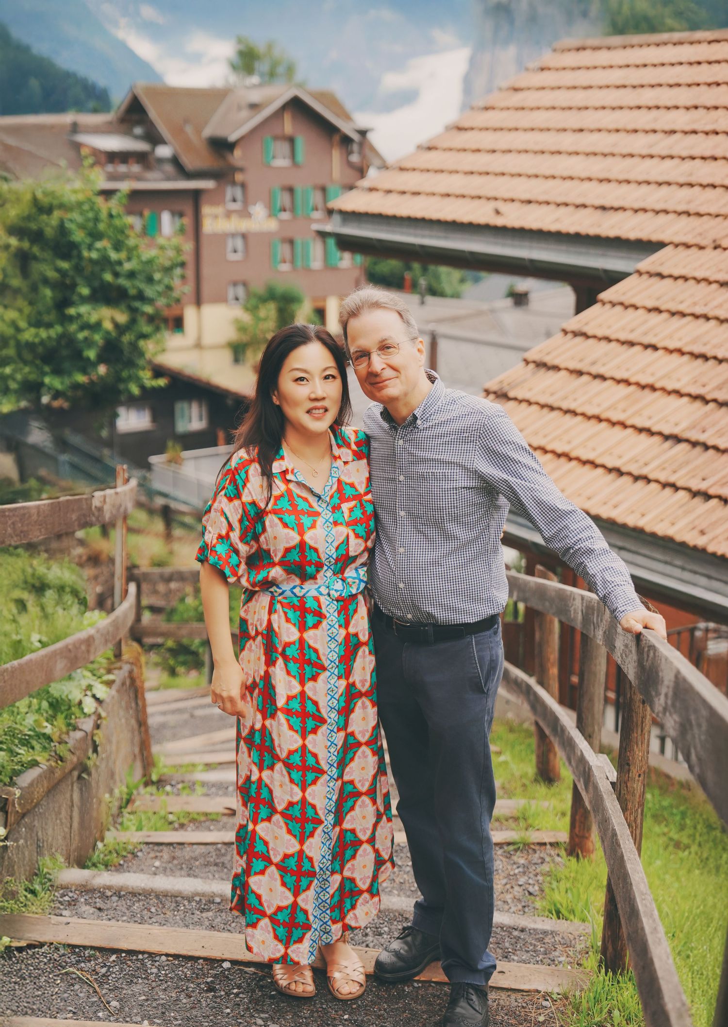 A couple in colorful attire poses on wooden steps with a lush Swiss village landscape behind them.