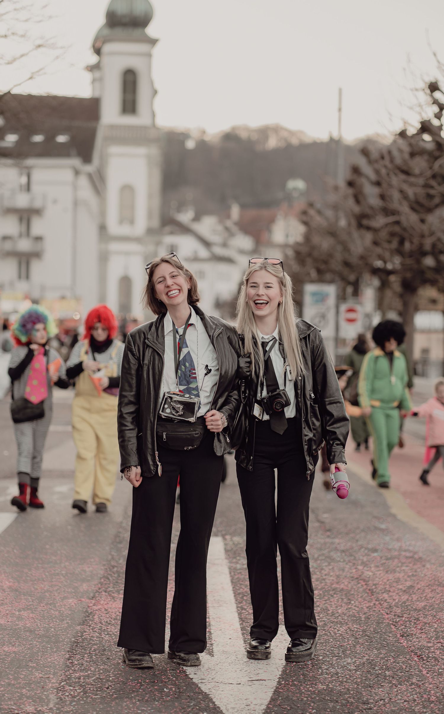 Two women in matching black leather jackets and wide-leg pants laugh while walking through a colorful carnival street parade.