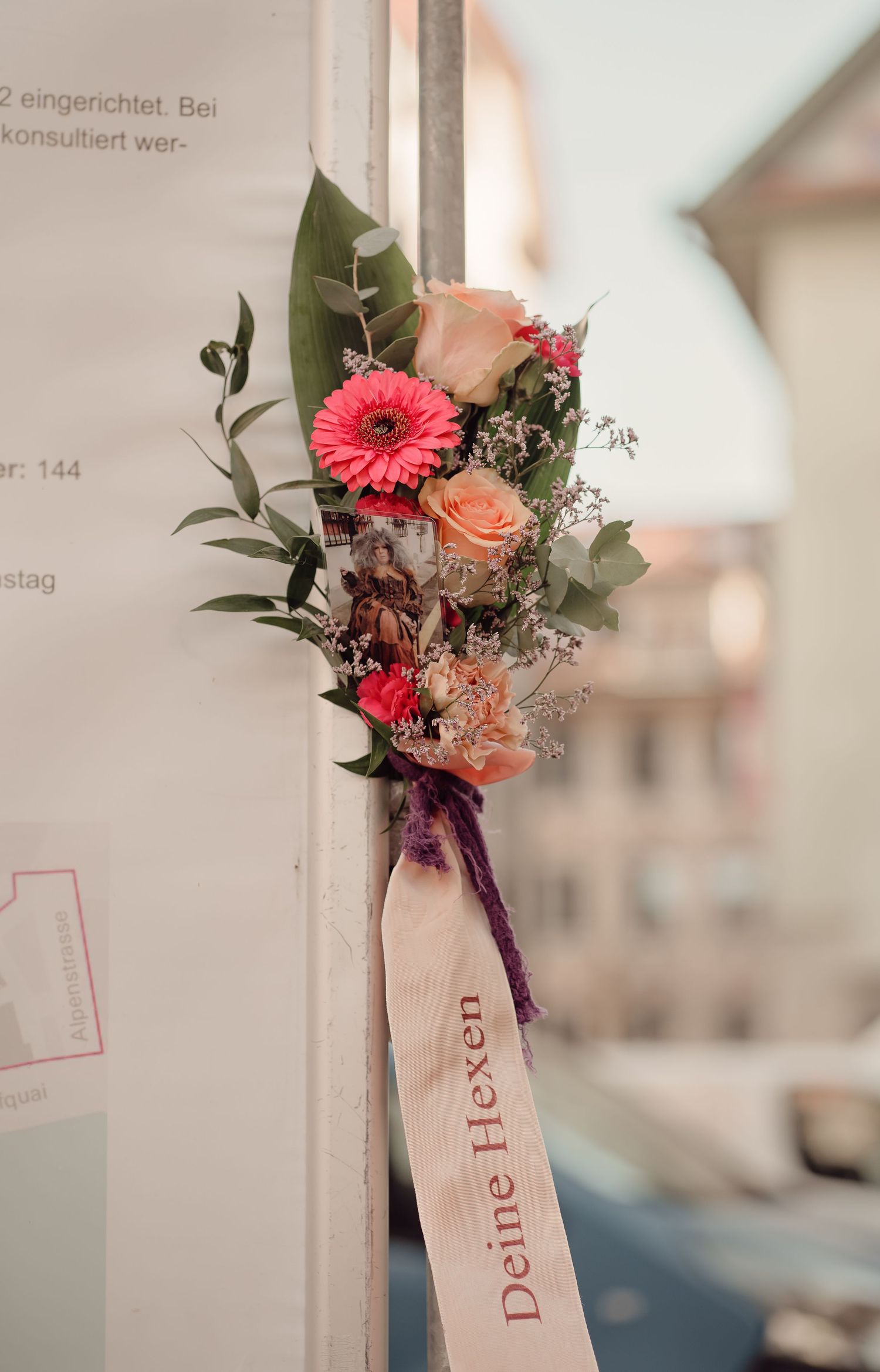 Small floral bouquet with pink roses and gerbera attached to a wall with a ribbon reading Deine Hexen.