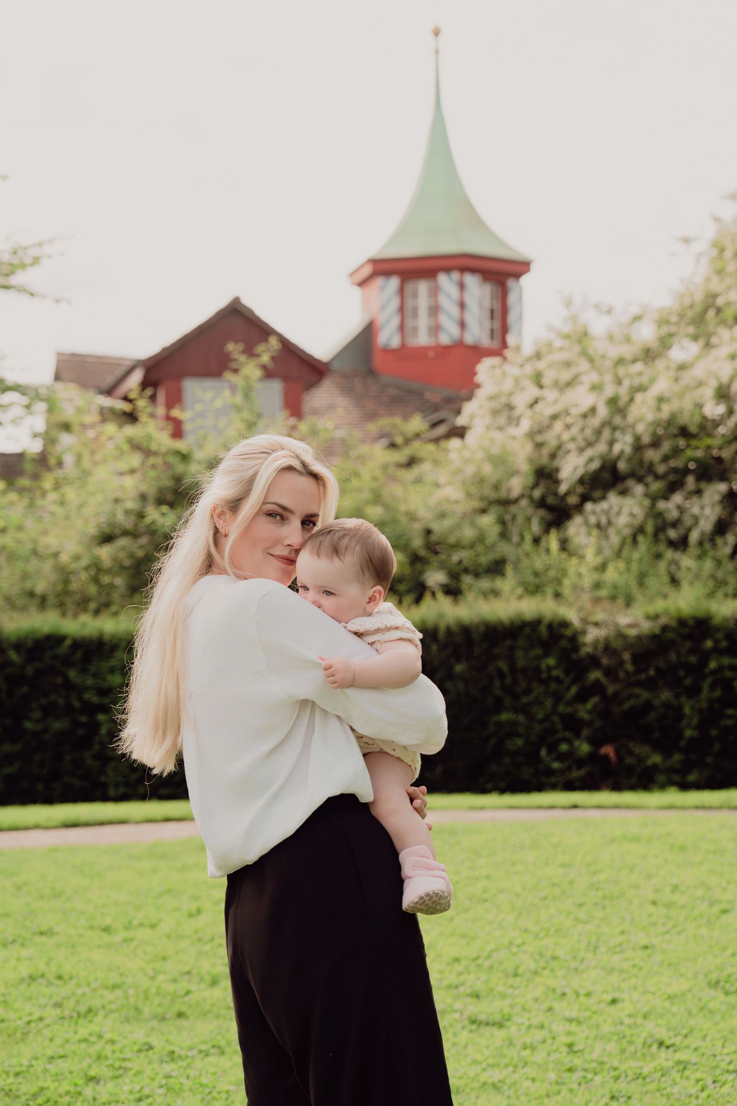 Blonde woman in white sweater holding a baby in a lush green park with a red tower building backdrop.