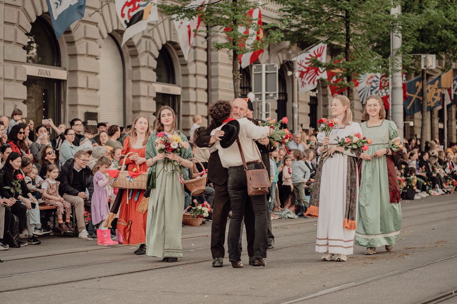 Participants in historical costumes embrace and carry flowers during a festive outdoor parade.