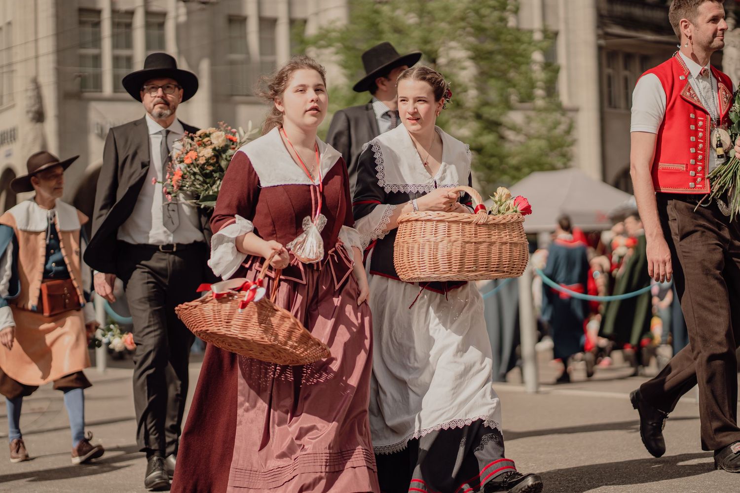 Women in traditional historical dresses carry flower baskets while marching in a cultural street parade.