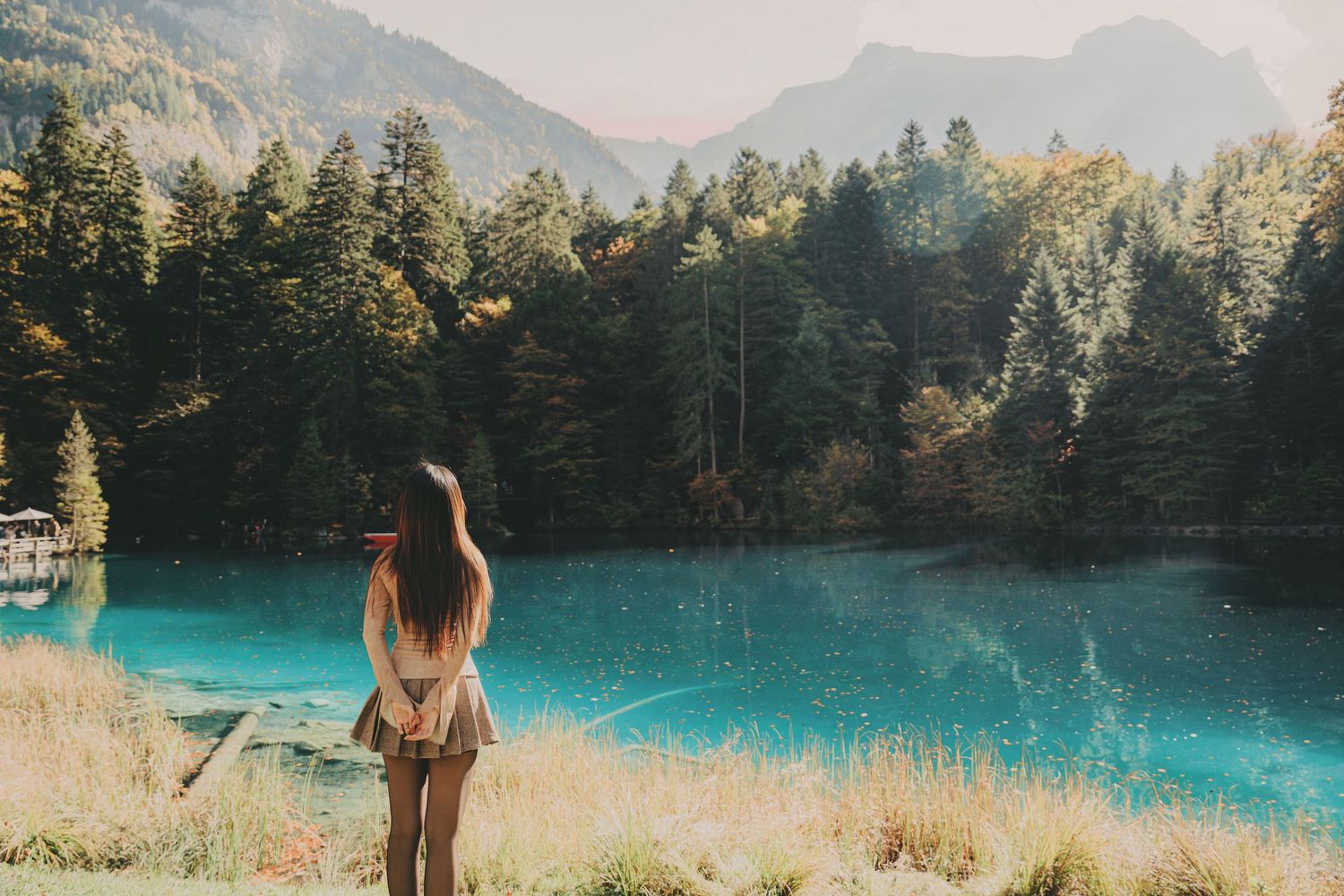 Woman with long hair standing by vivid turquoise lake surrounded by pine forest.