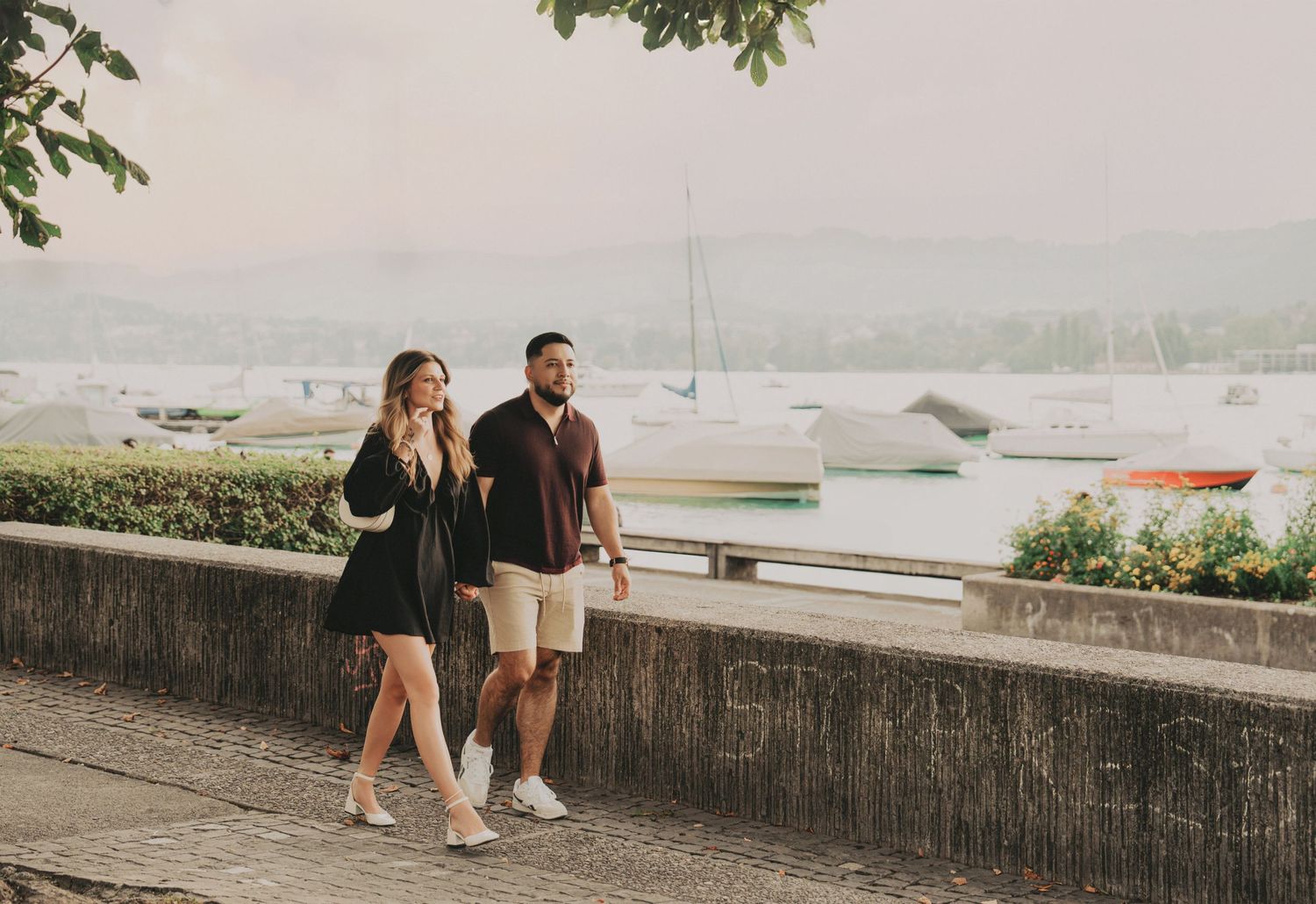 Couple walking hand in hand along a lakeside promenade with sailboats visible in the background.