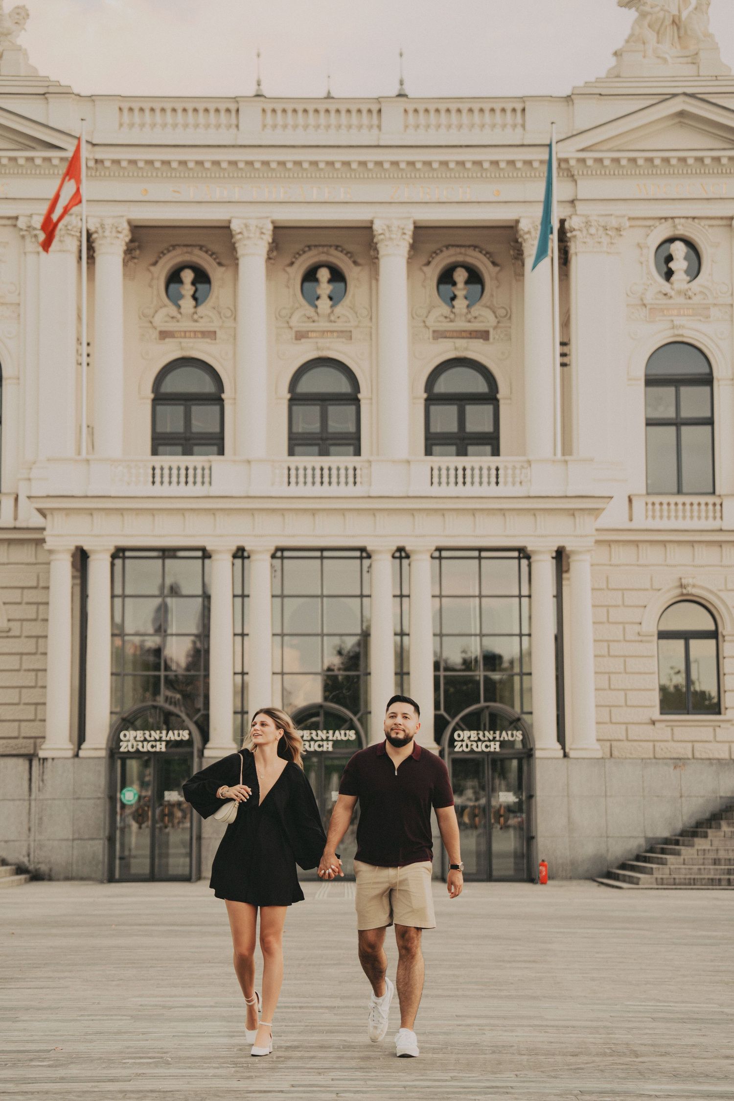Couple walking hand in hand toward the camera in front of a neoclassical European opera house.