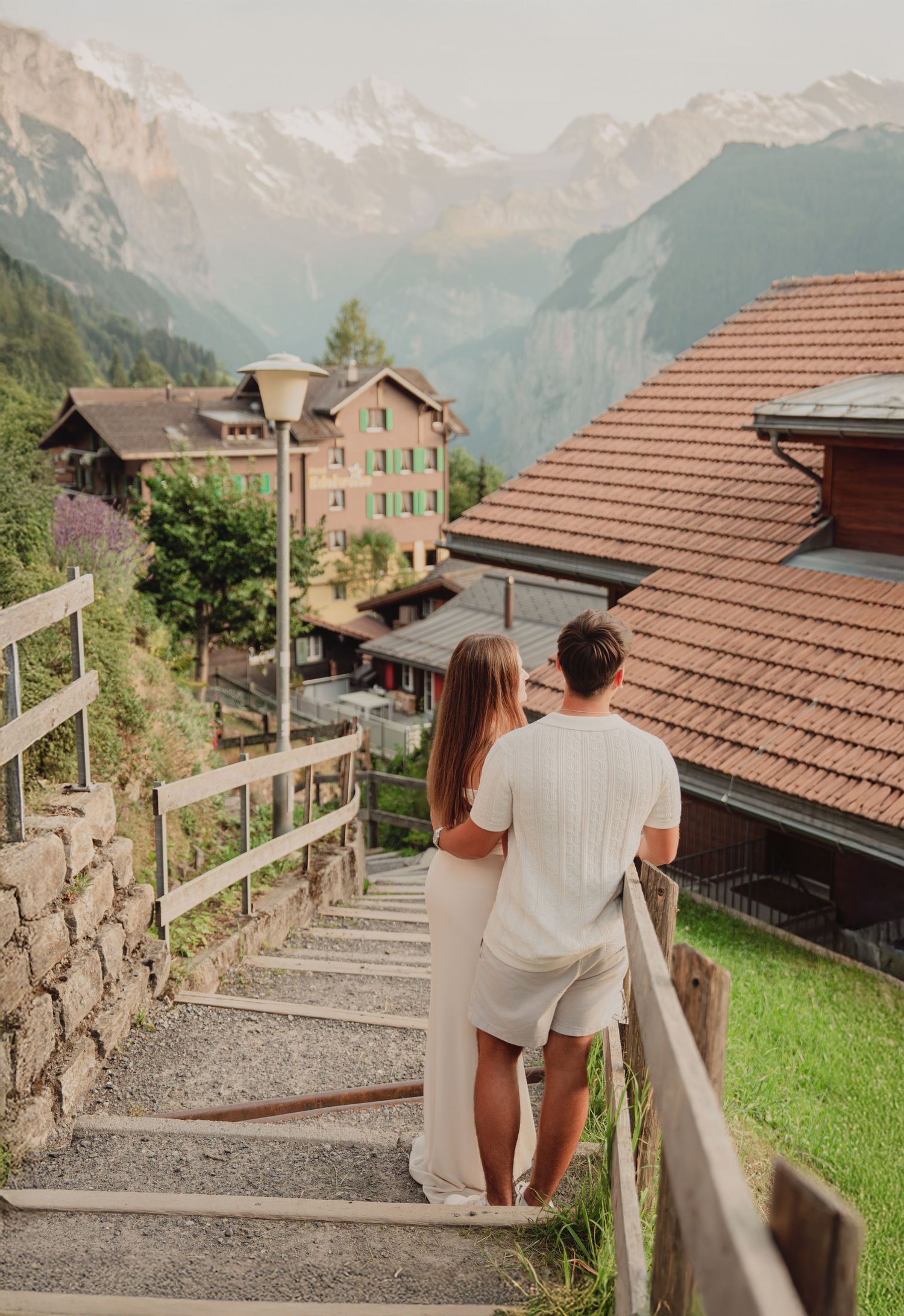 Couple standing together on stone steps admiring stunning Swiss Alpine village scenery with snow-capped mountains.