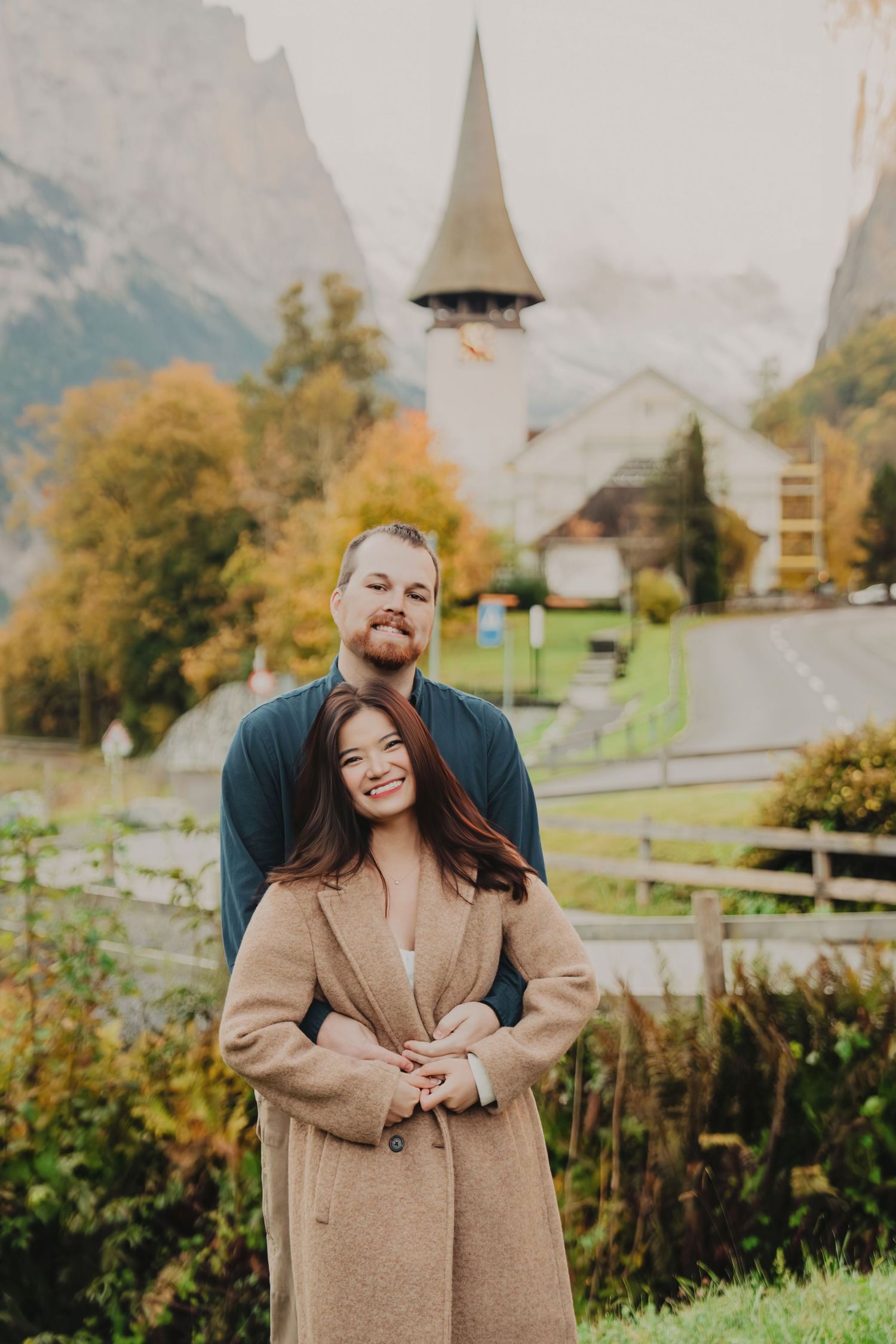 Couple posing warmly in front of a pointed church steeple surrounded by colorful autumn foliage in the Alps.