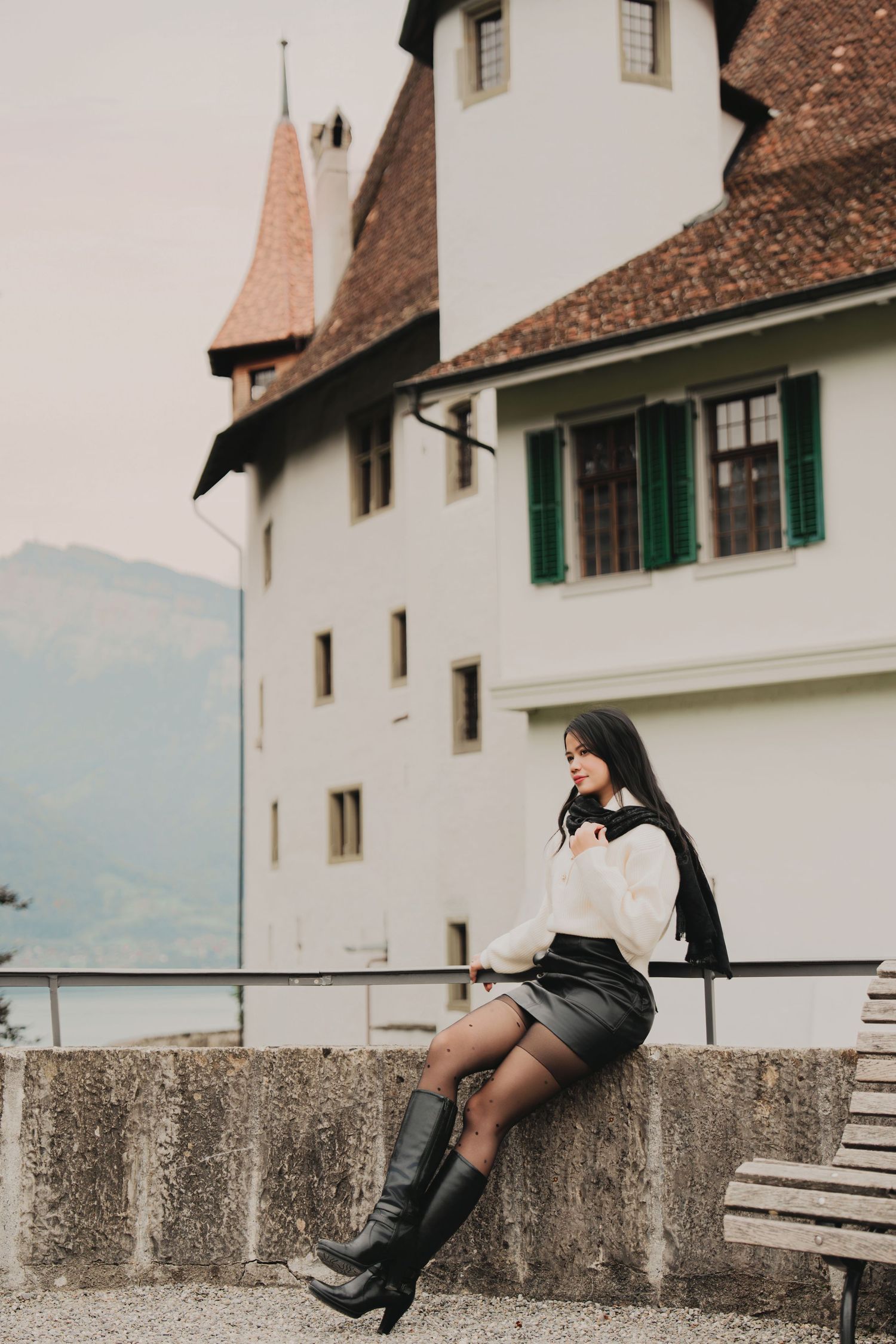 Elegant woman in white sweater sits on stone wall beside historic Swiss castle with green shutters.