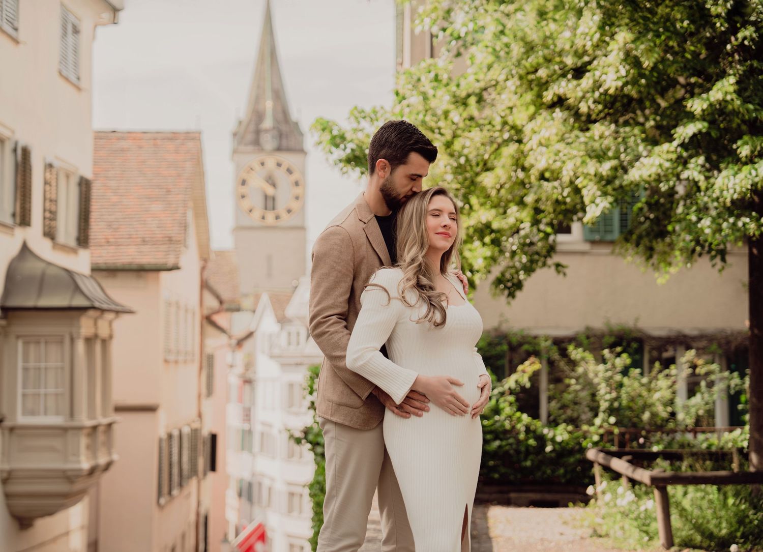 Couple shares tender embrace during maternity photoshoot in historic Zurich with St. Peter church tower backdrop.
