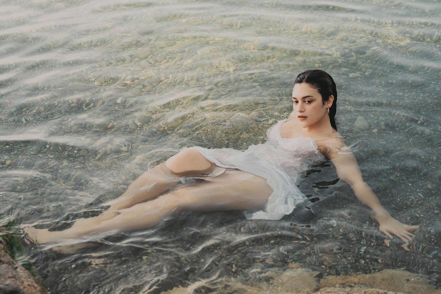 Woman in wet white dress reclining in shallow rocky water, gazing into distance at golden hour.
