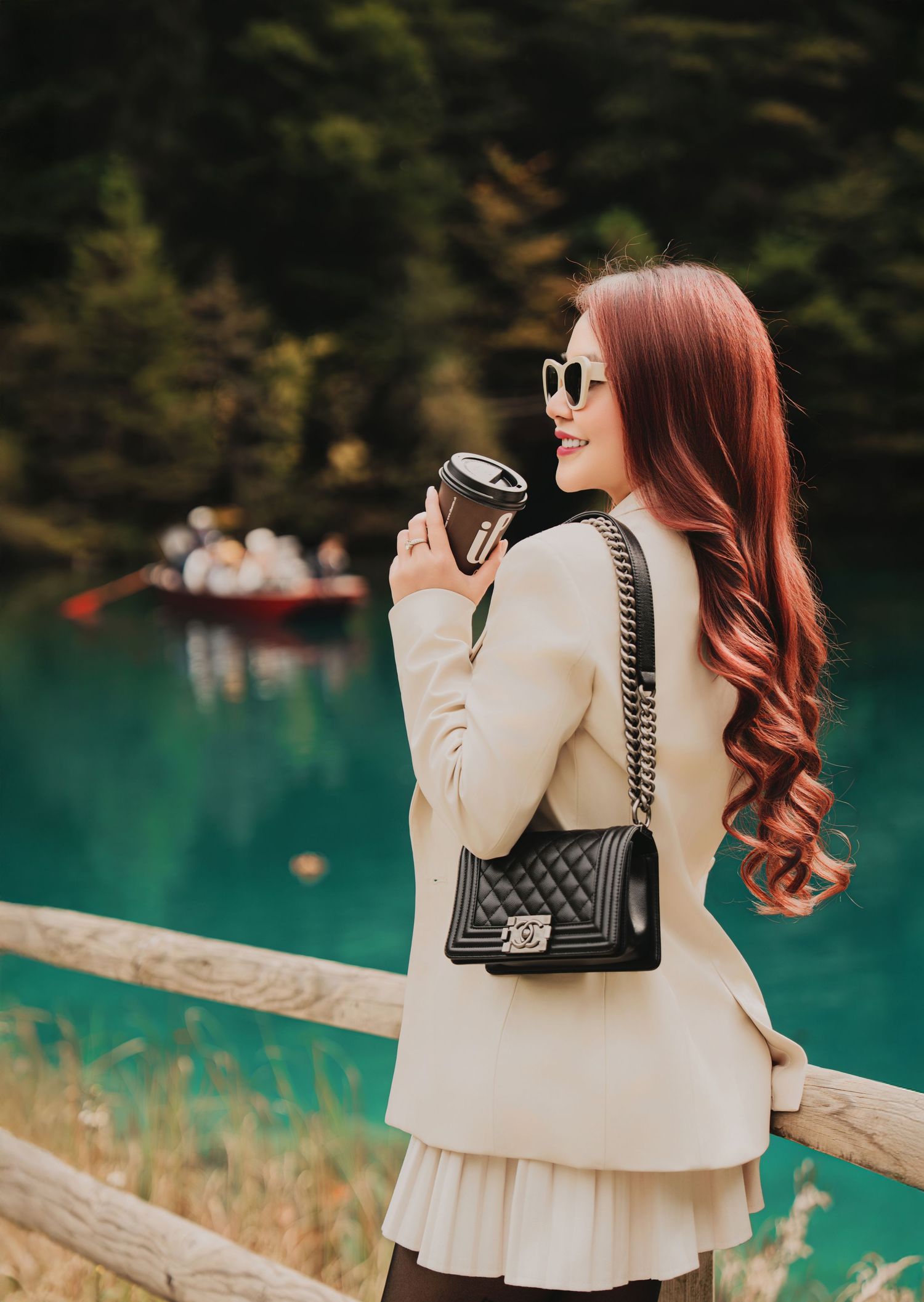 Elegant woman with Chanel bag and coffee cup smiling by turquoise lake with rowing boat.