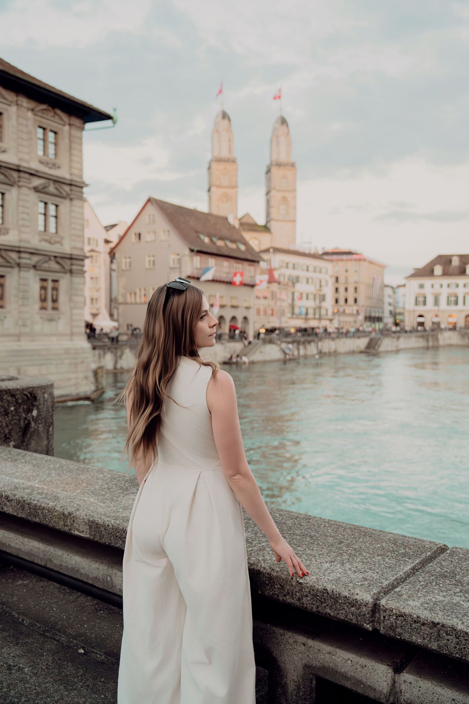 Young woman in white jumpsuit poses by Zurich river with Grossmünster cathedral twin towers in background.