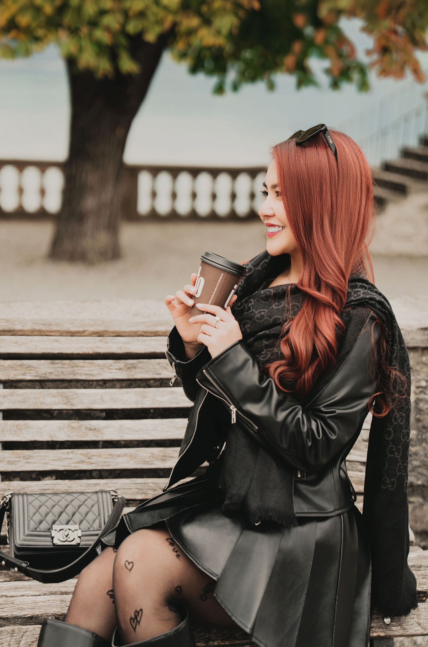 Smiling red-haired woman in black leather outfit enjoys coffee on park bench with autumn foliage behind.