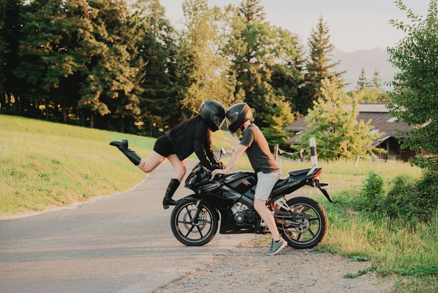 Couple sharing a kiss on a black sport motorcycle parked on a scenic road with green trees.