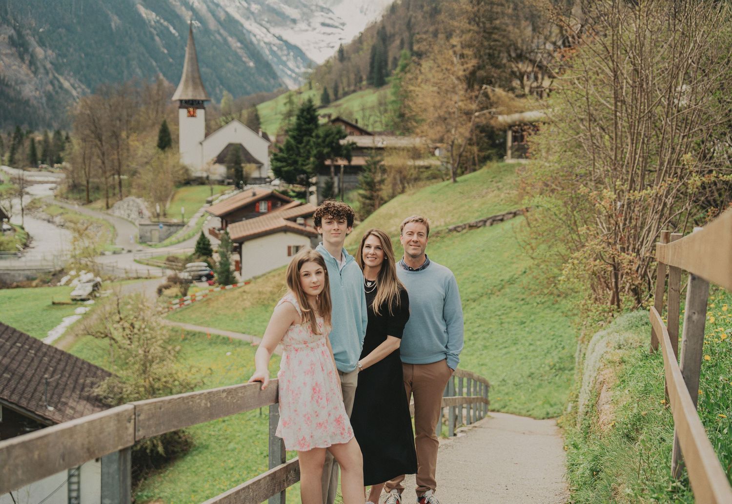Family smiling on a wooden bridge overlooking a scenic Swiss Alpine village and church.
