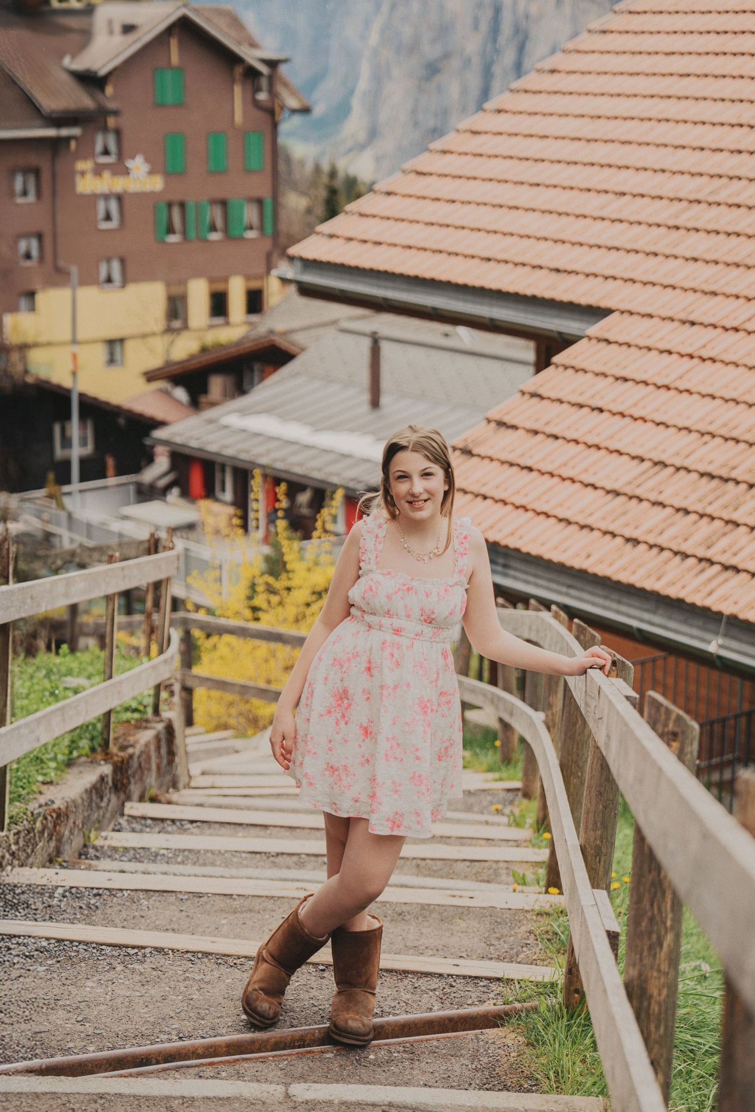 Smiling woman in floral dress and brown boots standing on wooden staircase in Swiss mountain village.