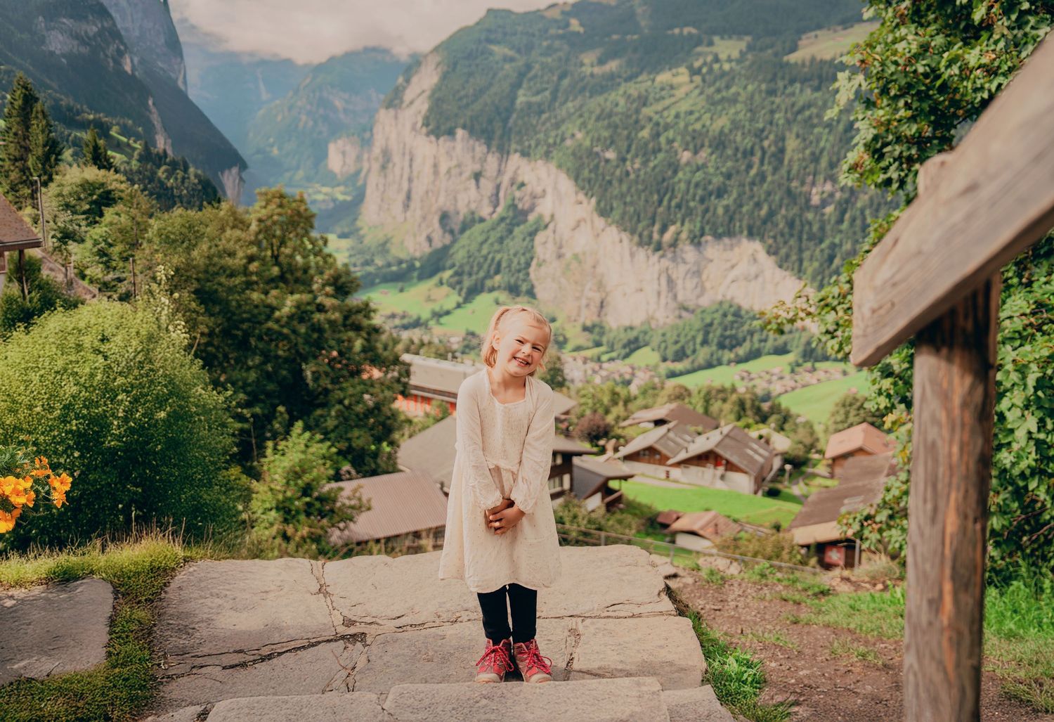 Young girl in cream dress with red boots standing on stone path in scenic Swiss mountain village.
