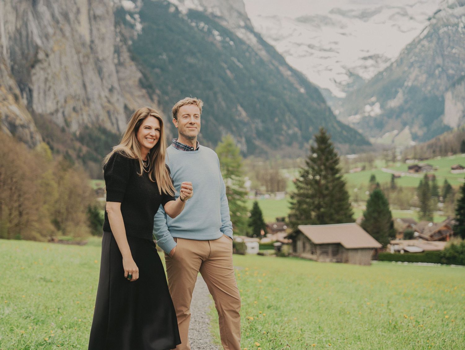 Couple smiling together on green meadow with stunning Swiss Alps and valley village behind them.