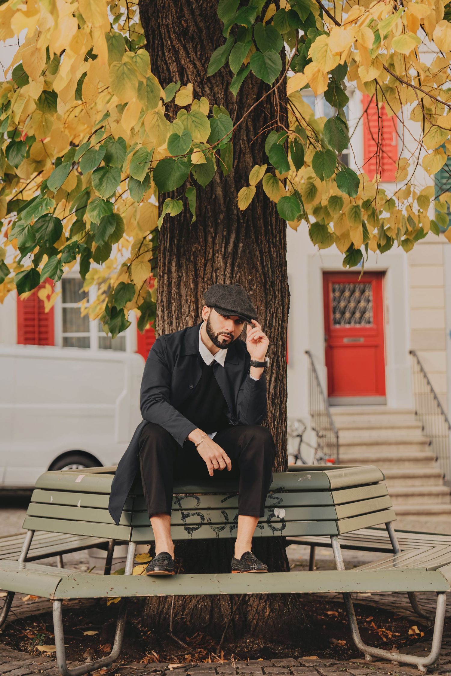 Bearded man in dark coat and flat cap sitting pensively on bench under autumn yellow-green tree.