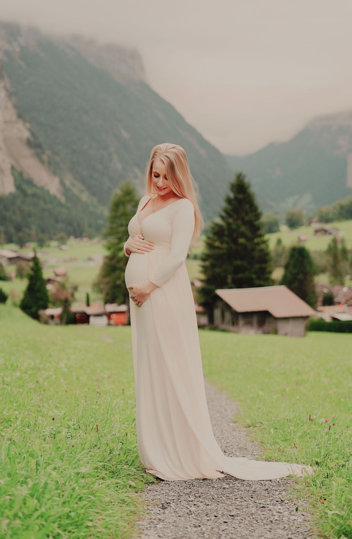 Pregnant woman in elegant cream maternity gown gently holding her belly while gazing down in a Swiss valley.