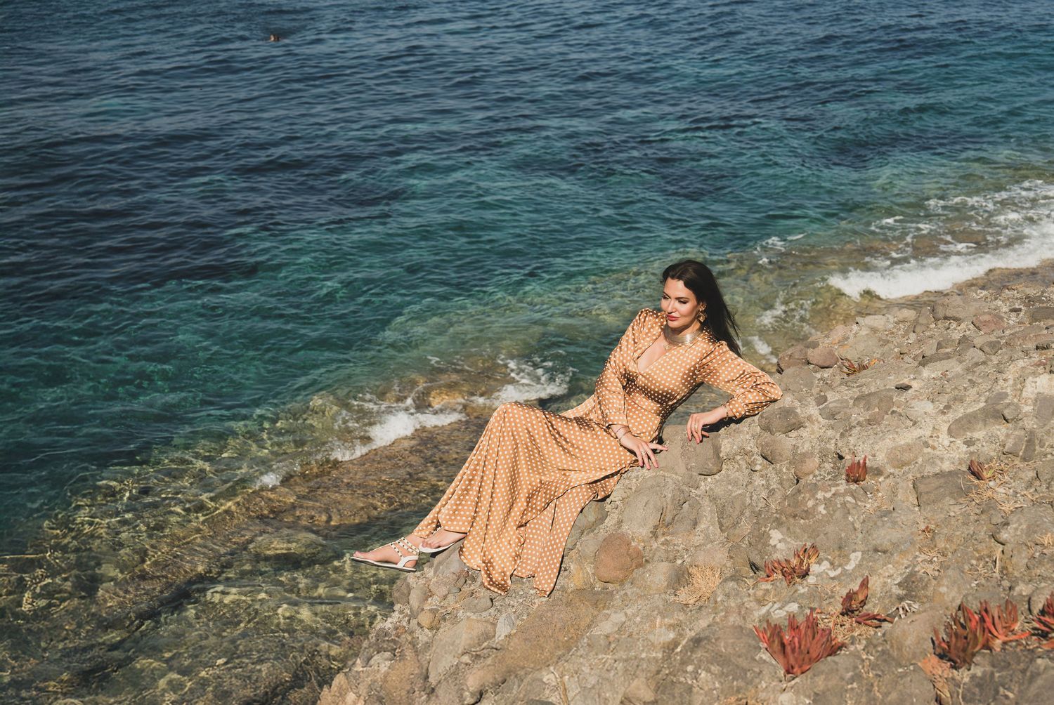 Woman in gold polka dot dress poses on rocky cliffside above stunning blue Mediterranean waters.