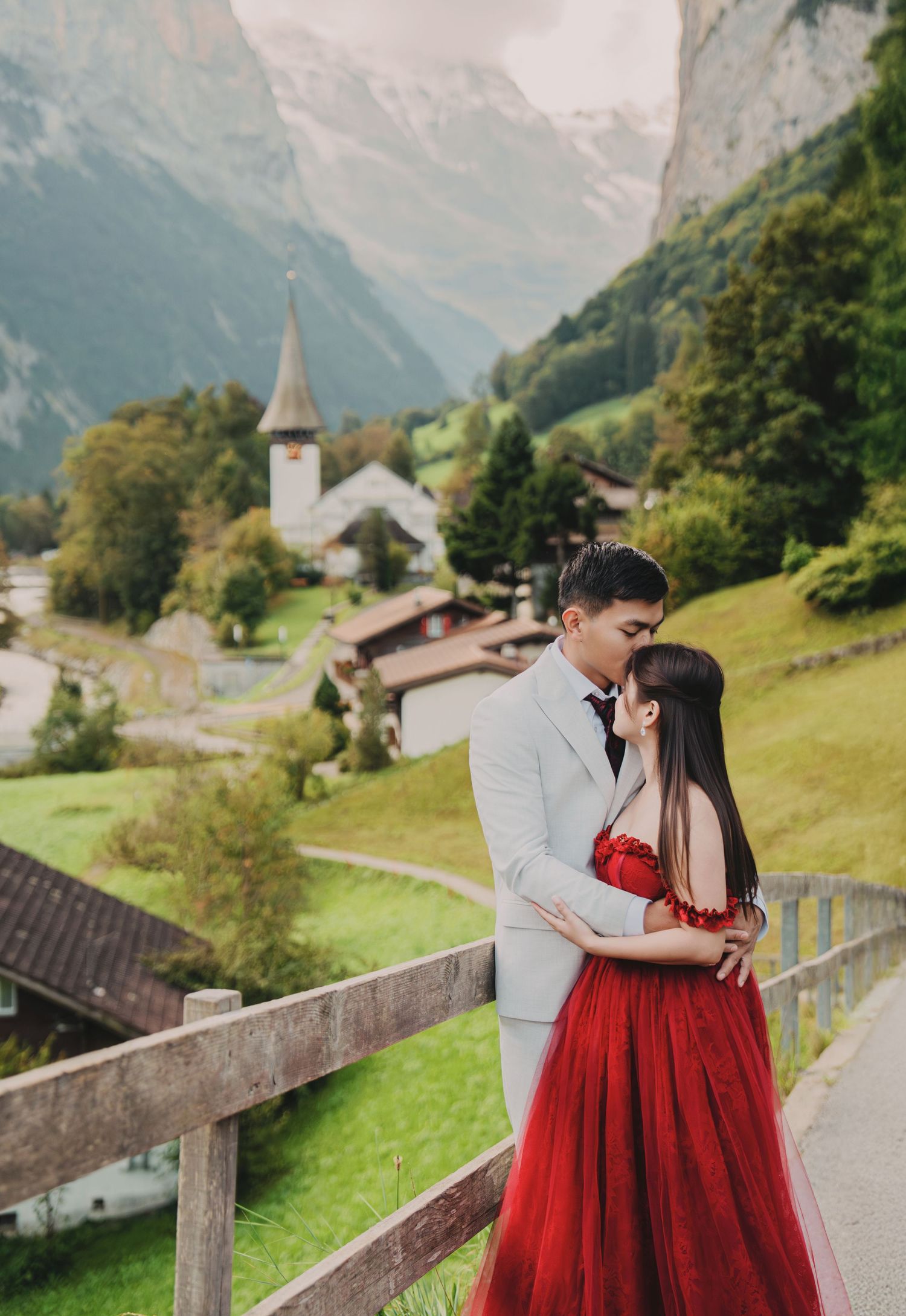 Couple sharing a tender kiss in red gown and grey suit with charming Swiss village and Alps behind.