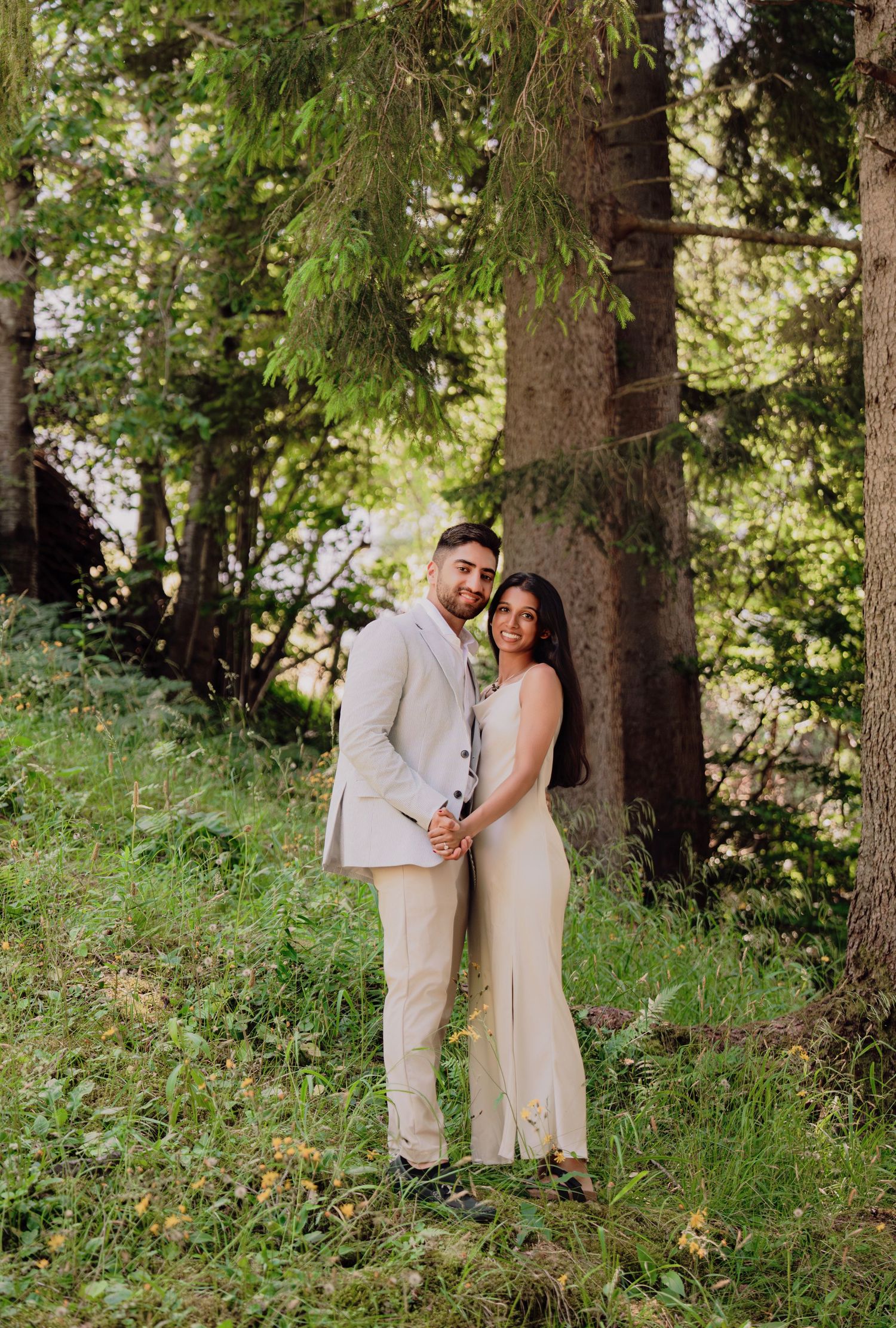 Bride and groom embracing in a lush green forest, wearing elegant cream wedding outfits.