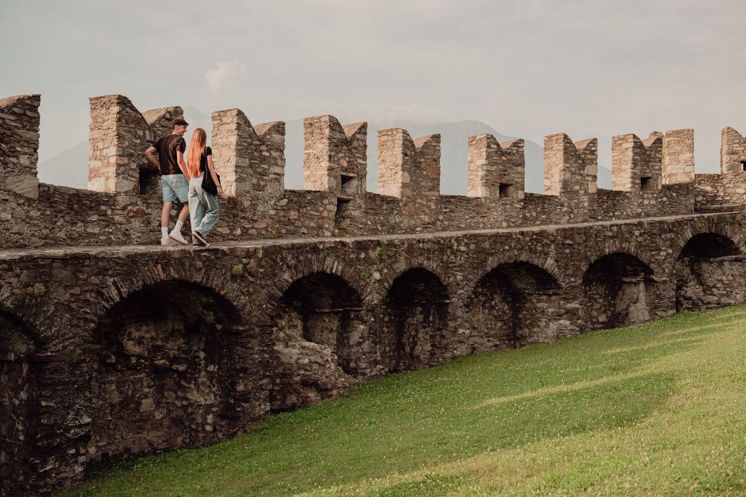 Couple walking along ancient stone castle battlements with arched walls and green grass below.