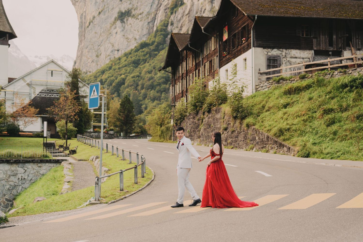 Couple holding hands crossing road in Swiss mountain village with dramatic cliff backdrop.