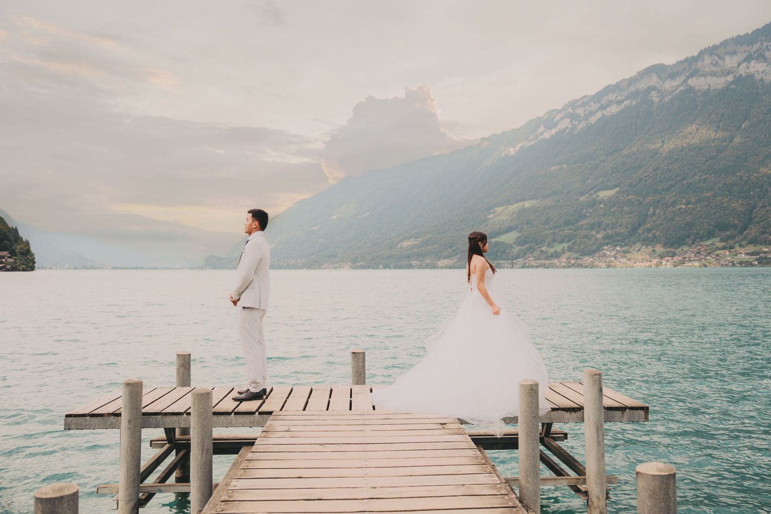 Bride and groom standing apart on wooden dock overlooking majestic alpine lake and dramatic mountains.