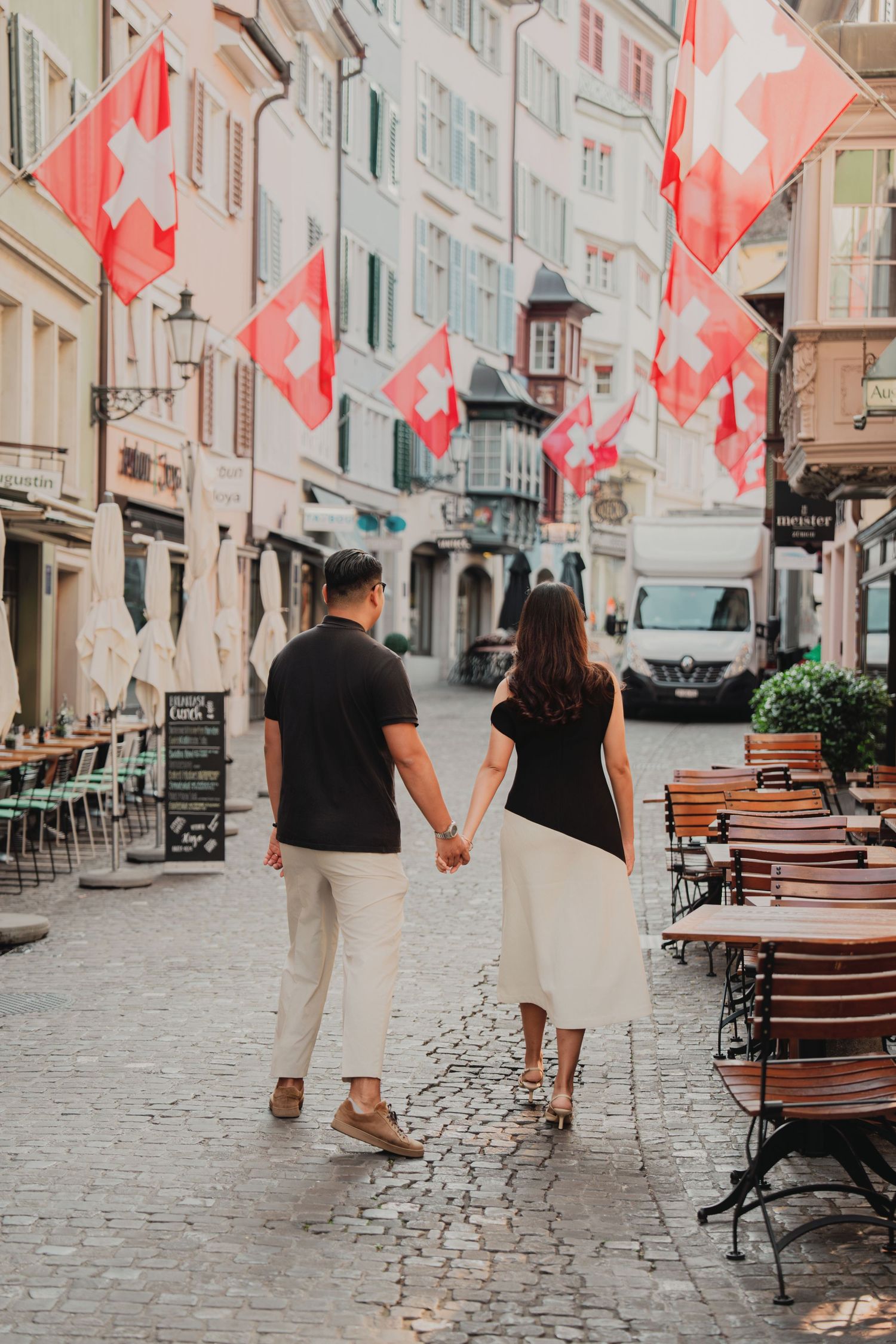 Couple walking hand in hand down a charming Swiss cobblestone street lined with red and white flags.