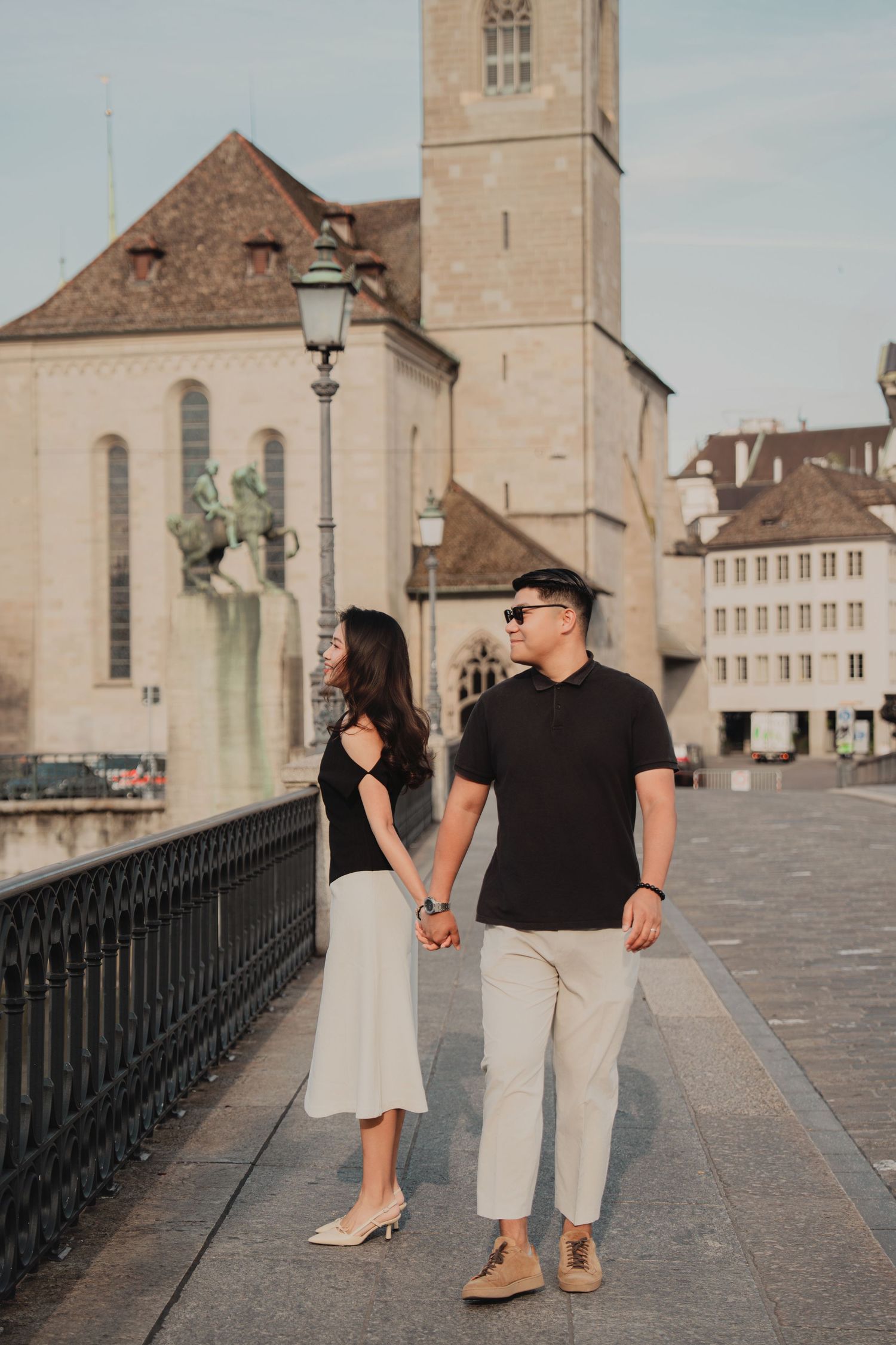 Couple walking hand in hand on a Zurich old town bridge with a church tower and buildings behind.