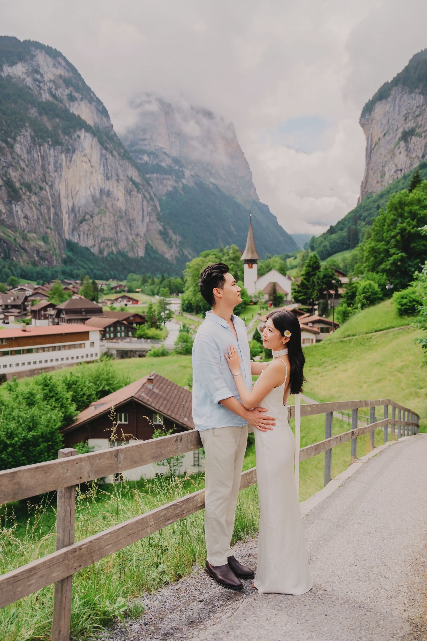 Couple sharing a tender moment on a wooden bridge with Lauterbrunnen valley and church in background.