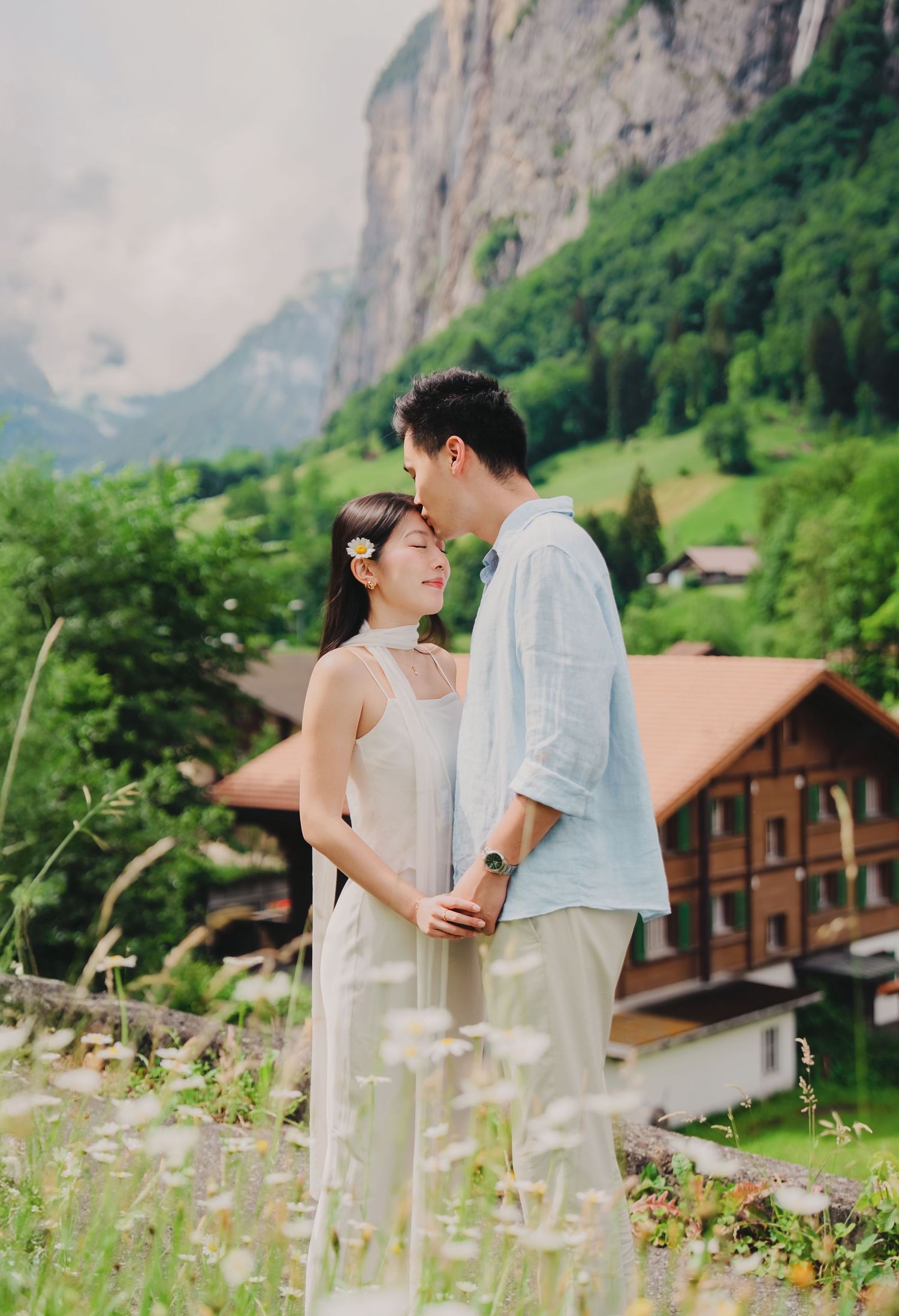 Couple embraces tenderly in front of a Swiss chalet with lush green mountains and wildflowers surrounding them.