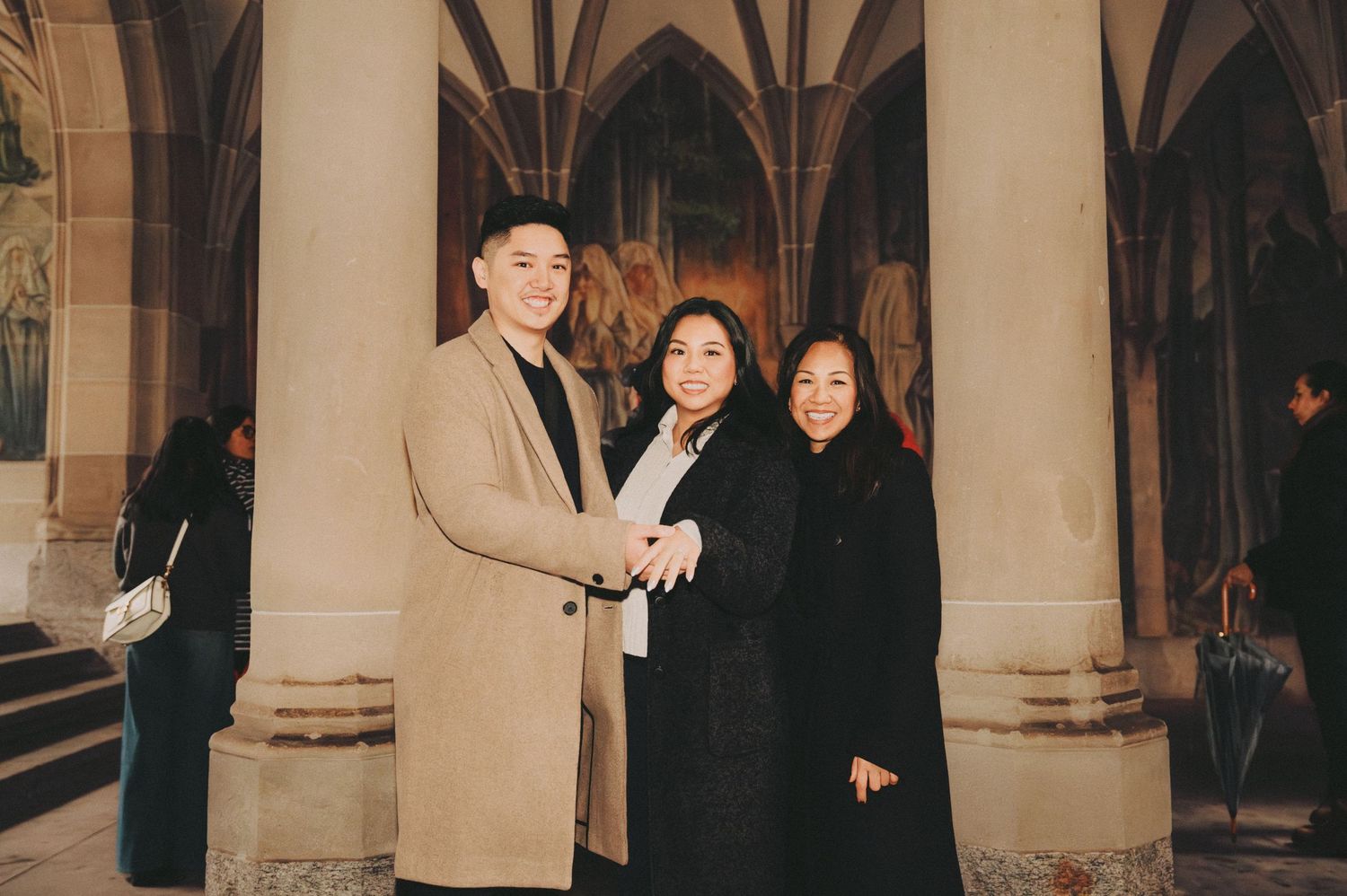 Three people smiling and posing together inside a gothic cathedral with ornate murals.