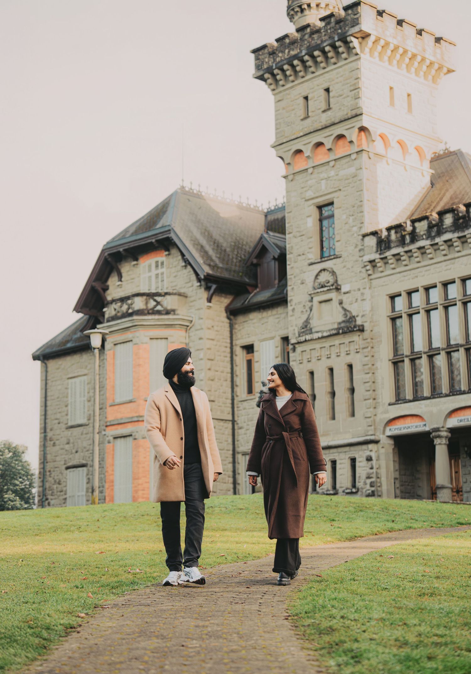 Engaged couple holding hands and walking along a path in front of a grand stone castle.