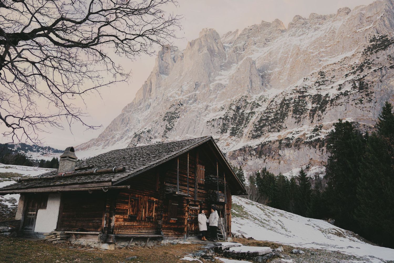 Couple standing before rustic alpine wooden chalet with massive snow-dusted rocky mountain in background.