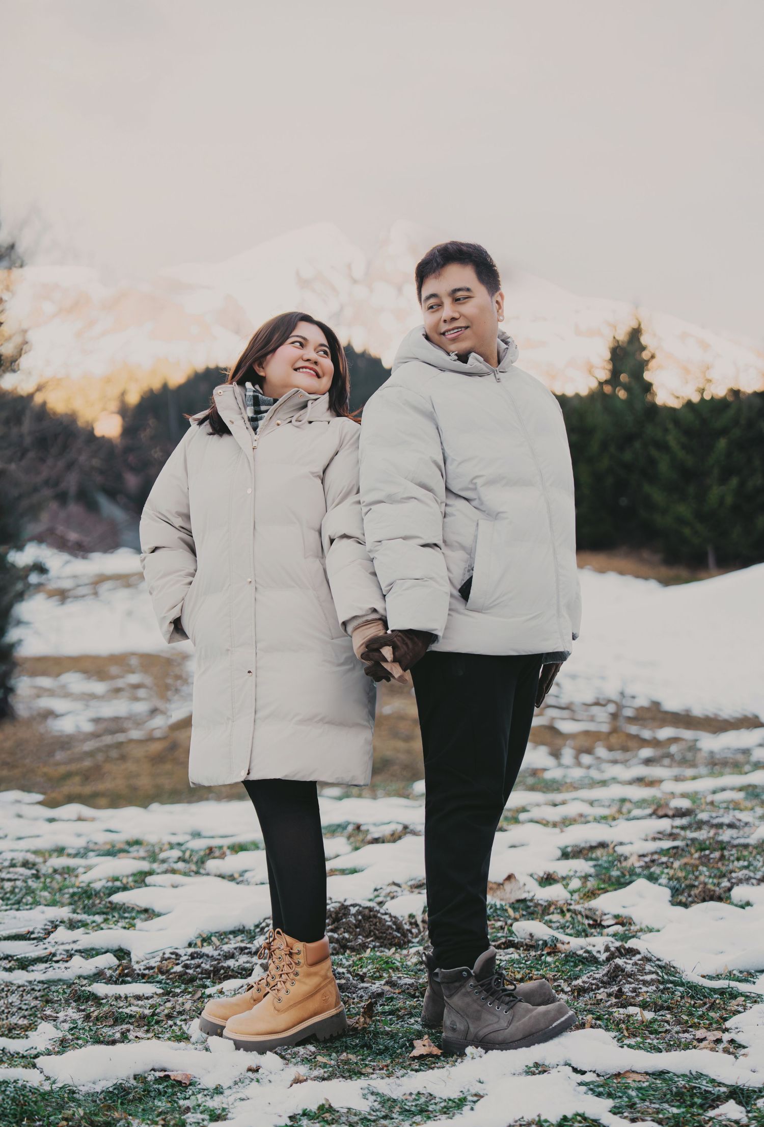 Happy couple holding hands in matching oversized white puffer coats with snow-capped mountain behind.