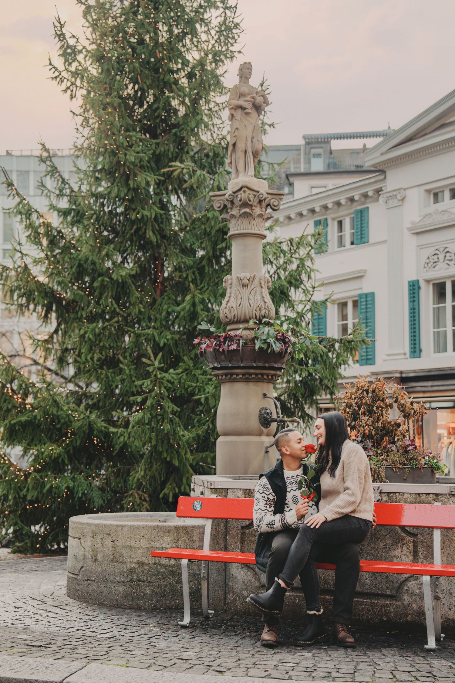 Couple sharing a romantic moment on a red bench near a European town fountain.