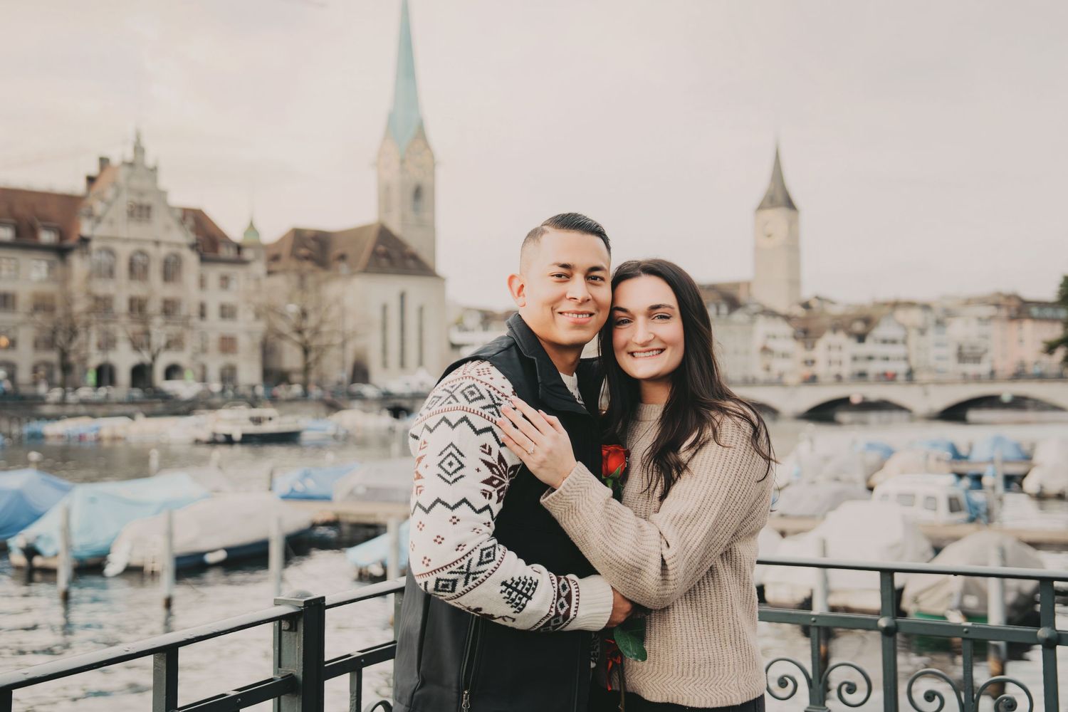Smiling couple embracing on bridge with Zurich harbor and church spires behind them.