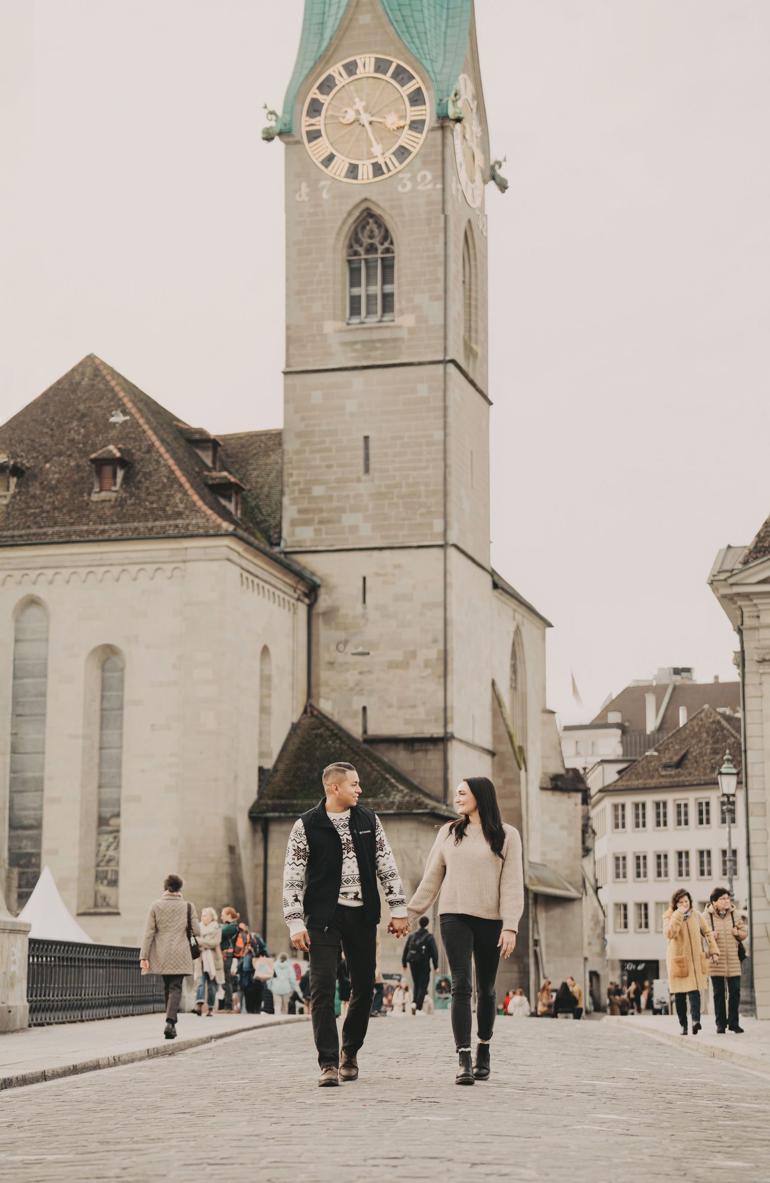 Couple holding hands walking in Zurich city square with Fraumunster church clock tower behind them.