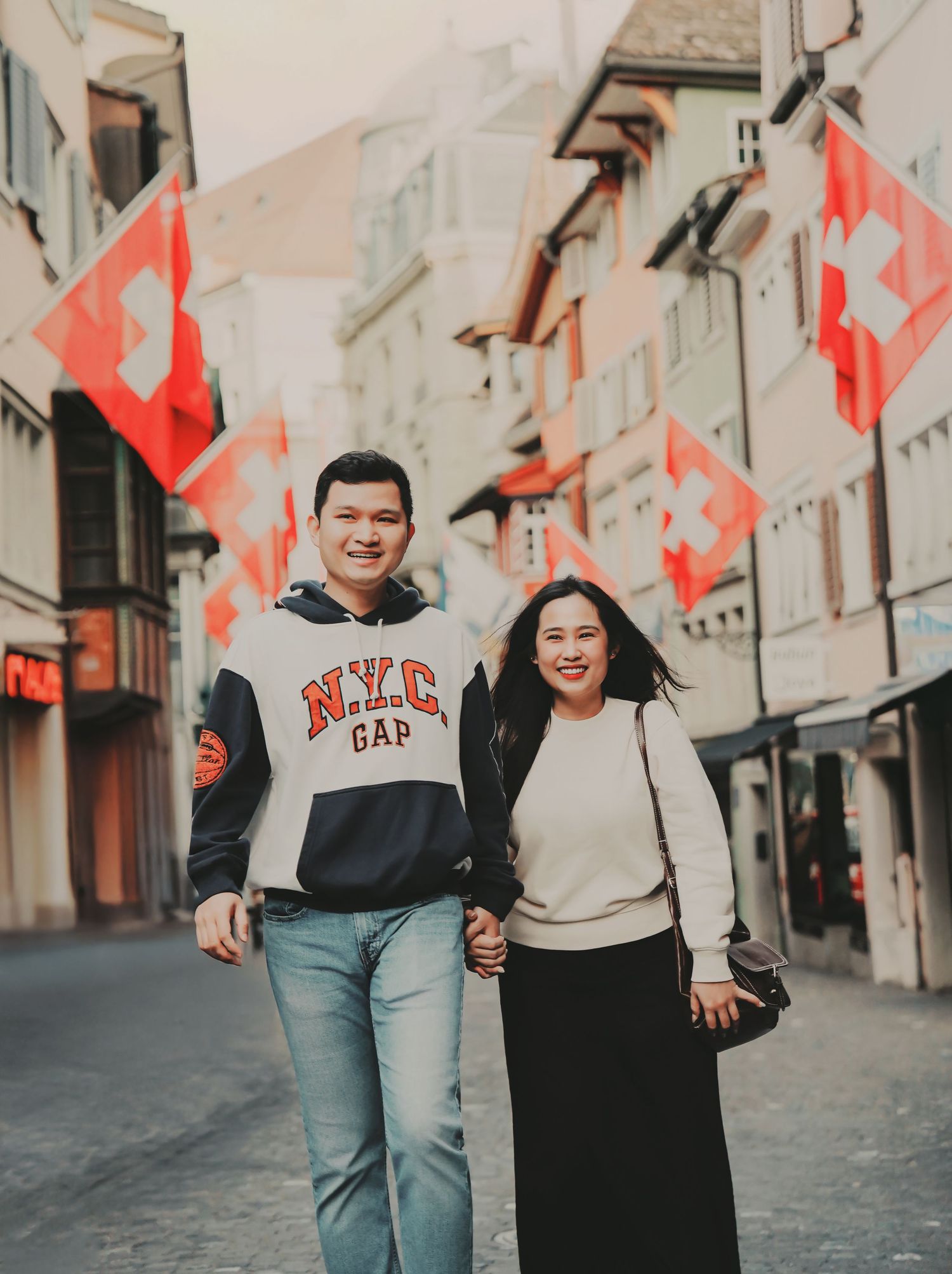 Smiling couple walking hand in hand down a Zurich street lined with Swiss flags on a cloudy day.