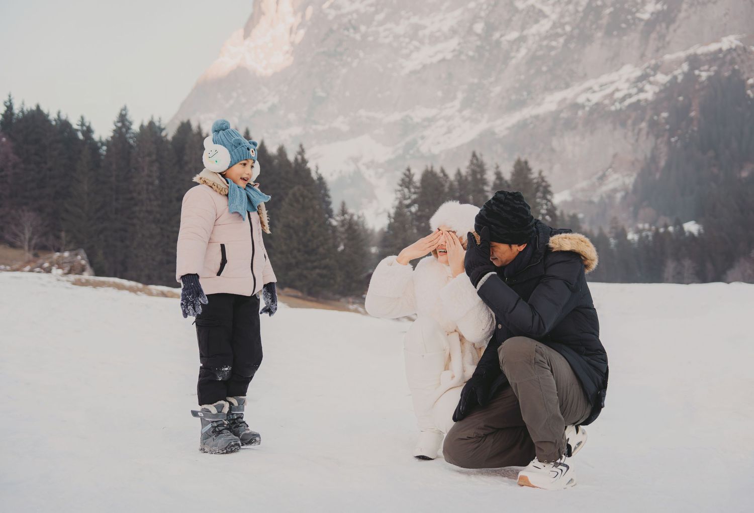 Man crouching to help young child build snowman on snowy field with mountains and pine trees behind them.