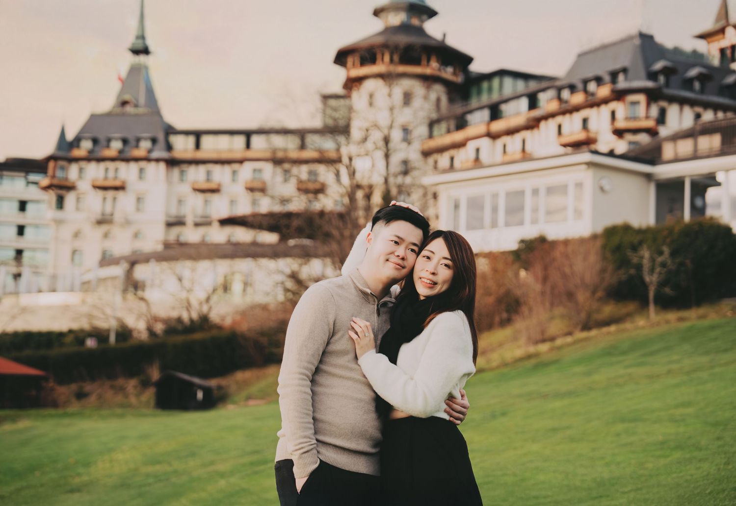 Loving couple posing closely together on a green lawn with an elegant castle building behind them.