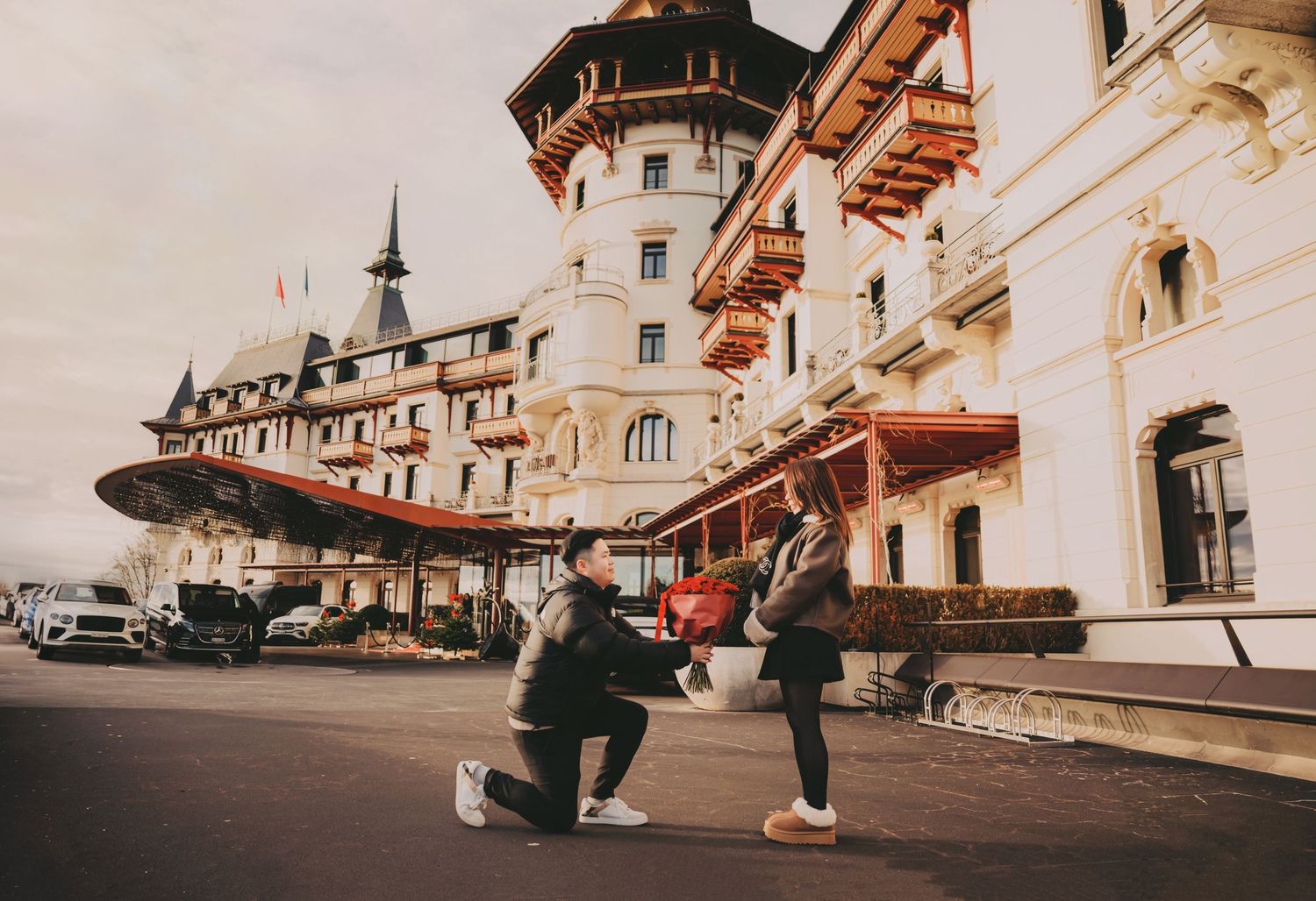 Man kneeling proposing to woman outside a grand European castle-style hotel building.