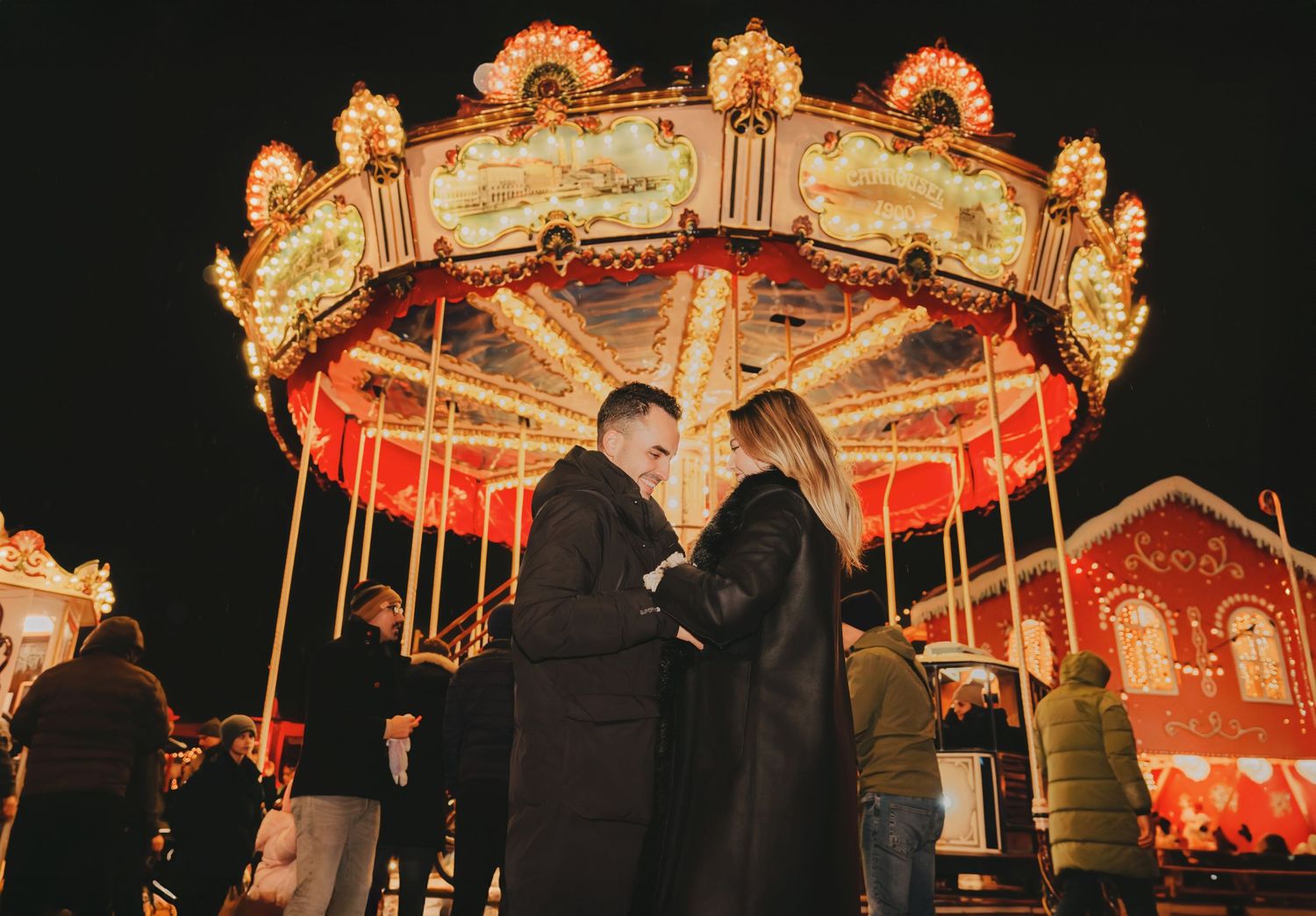 Romantic couple standing close together in front of a glowing carousel at a Christmas market at night.